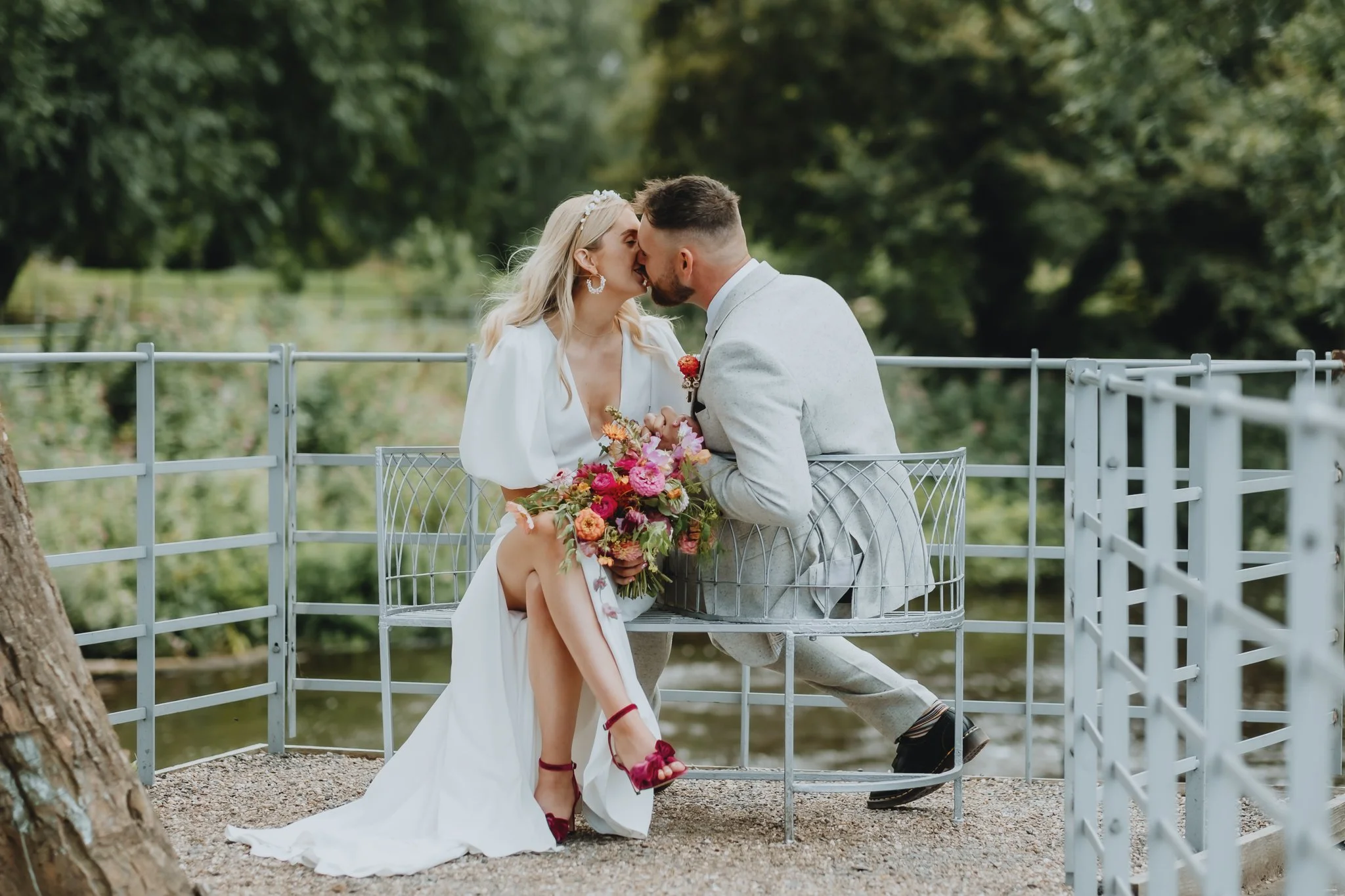 A newlywed couple sharing a kiss on a park bench near a pond, with the bride holding a colorful bouquet of flowers and wearing a white dress, the groom wearing a light gray suit.