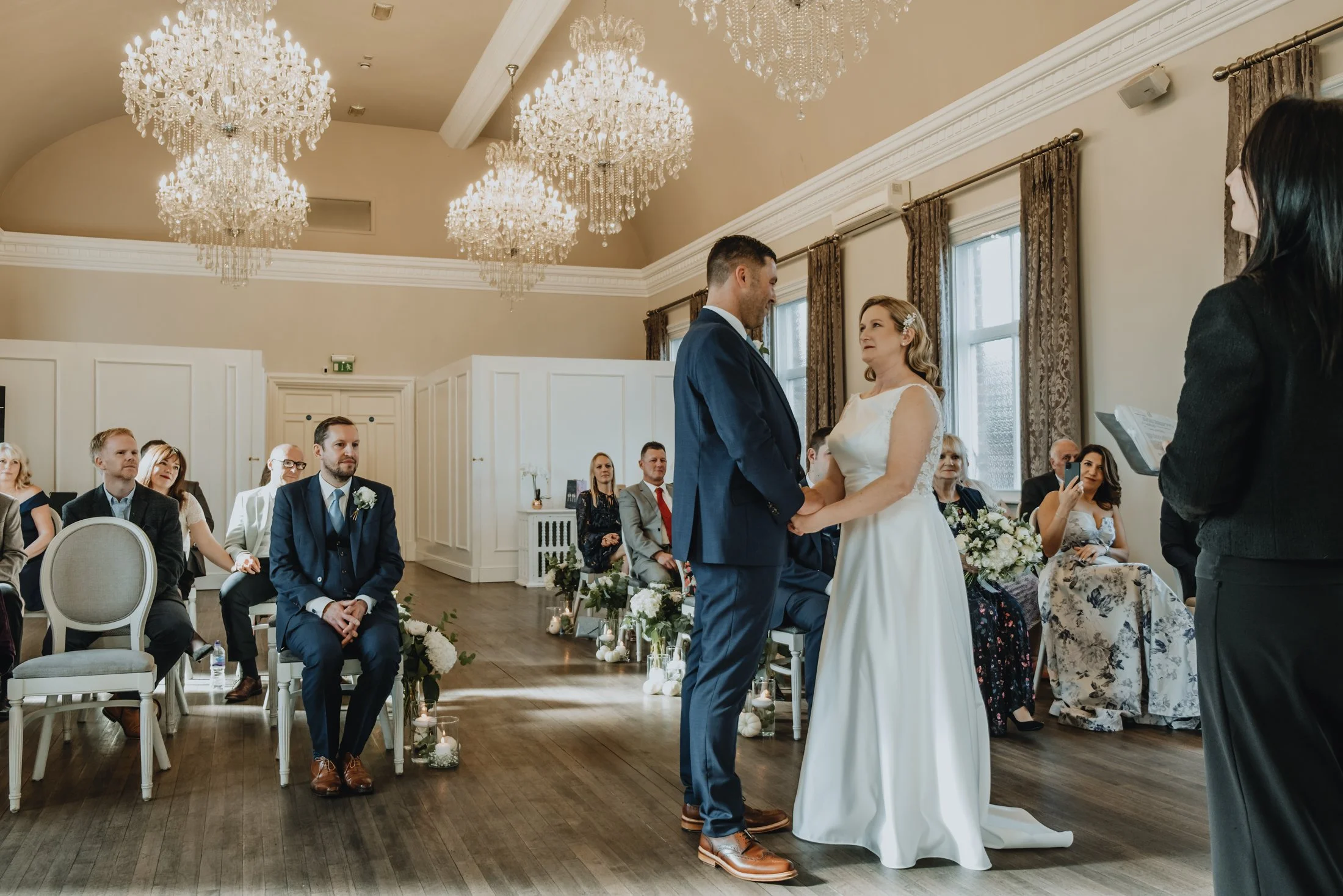 A wedding ceremony taking place in an elegant hall with chandeliers. The bride and groom are holding hands and looking at each other. Guests are seated on either side, watching the couple. The officiant is standing to the right, holding a book, durin