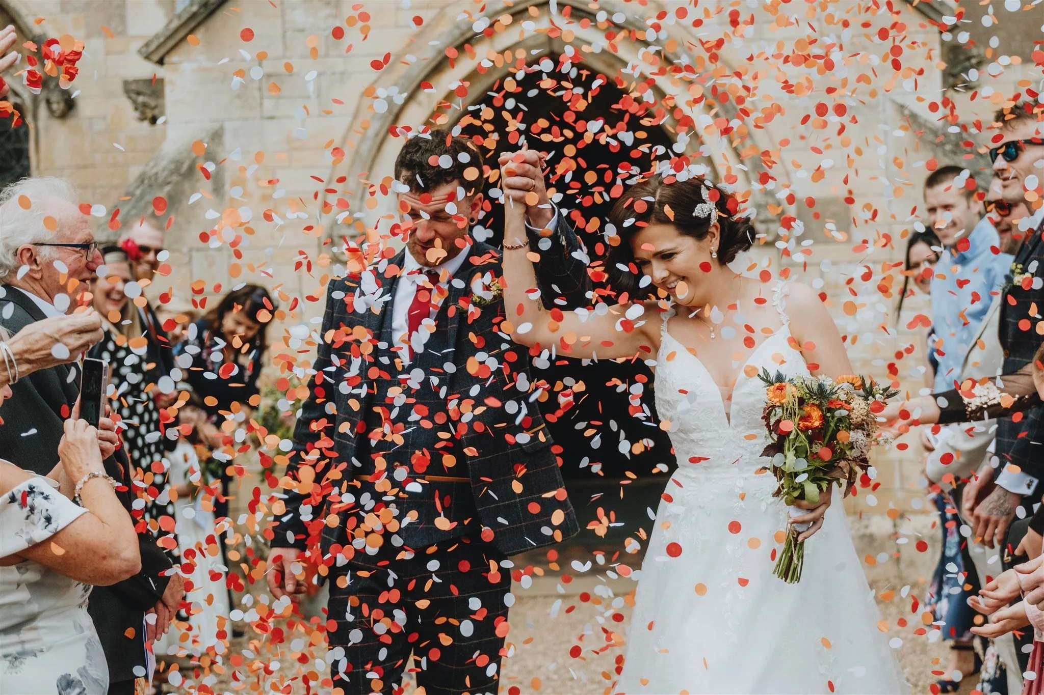 A bride and groom celebrating their wedding with confetti as they walk hand in hand, surrounded by friends and family outside a stone building.