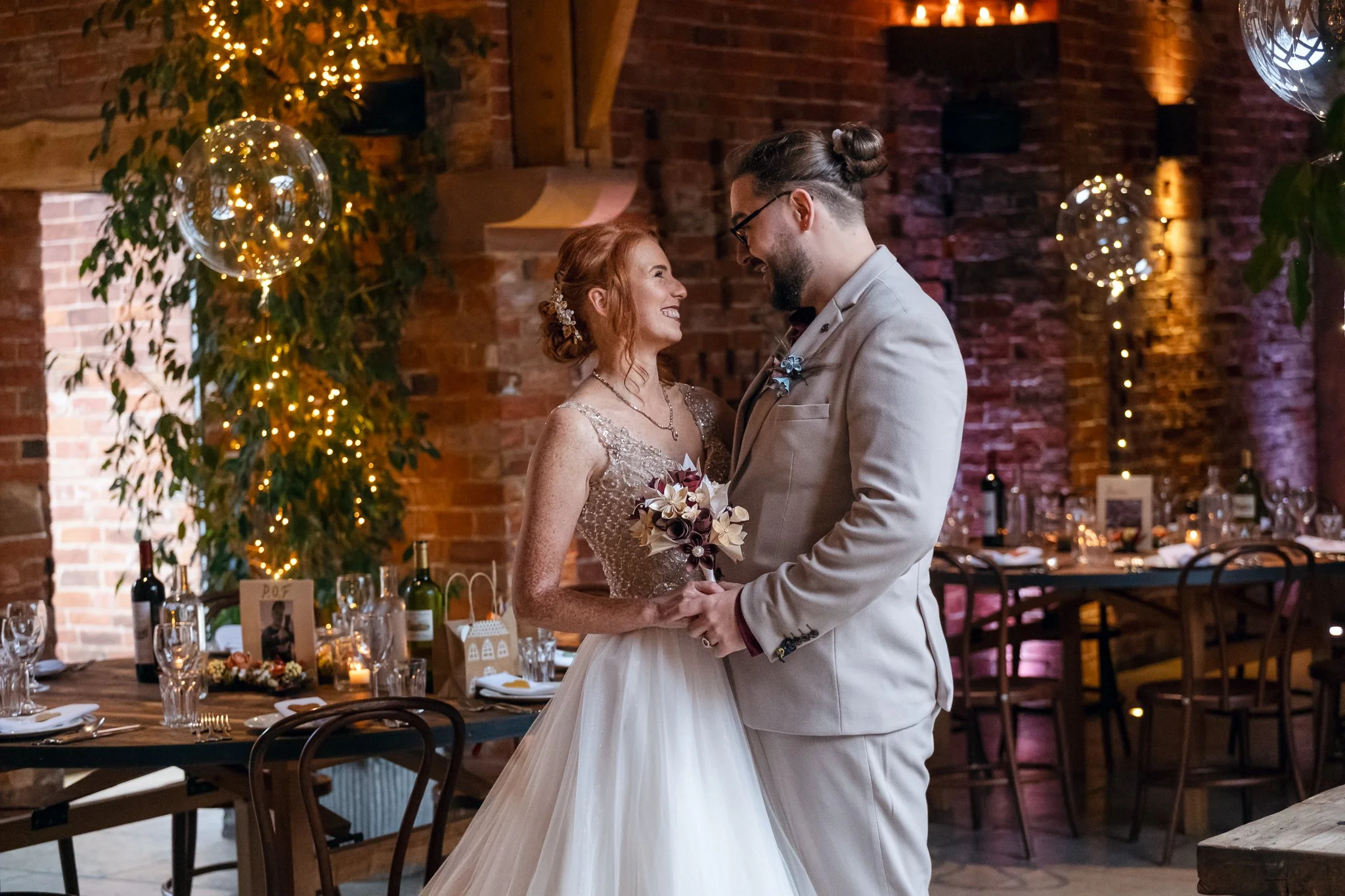 A bride and groom stand close, holding hands and looking at each other, smiling, in a decorated wedding venue with brick walls, fairy lights, and a wedding reception table in the background.