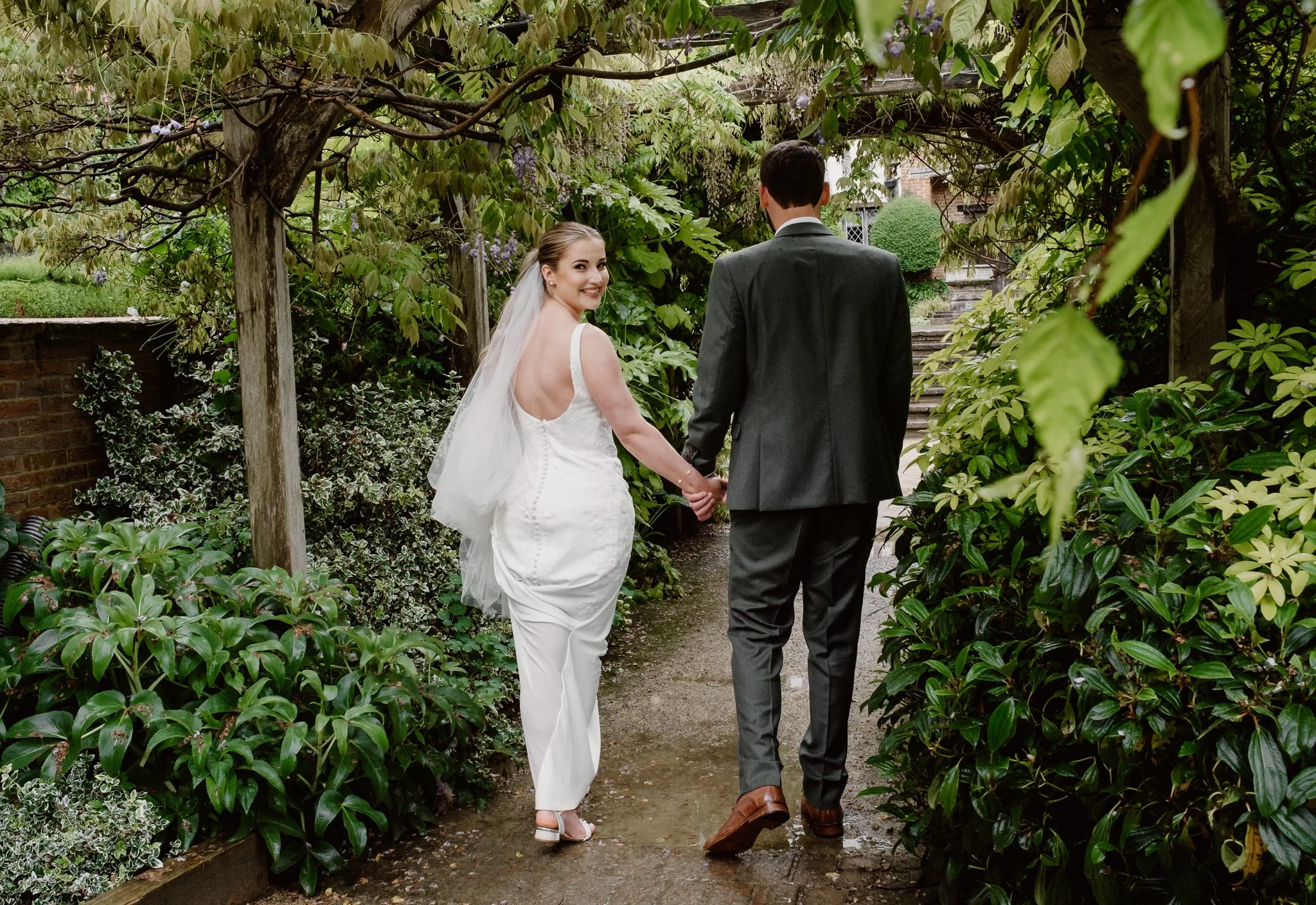 A bride and groom hold hands and walk away in a garden, with the bride smiling and looking back, dressed in a white gown and veil, and the groom in a dark suit.
