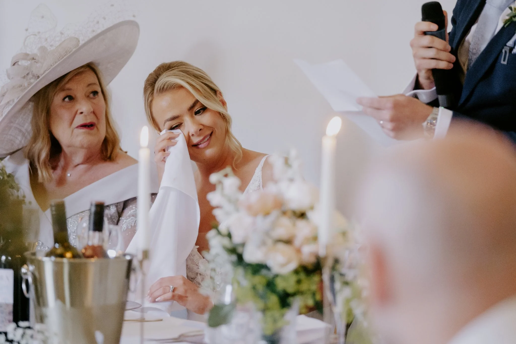 A woman in a wedding dress wiping tears, seated next to an older woman in a large hat, at a wedding reception with candles and flowers
