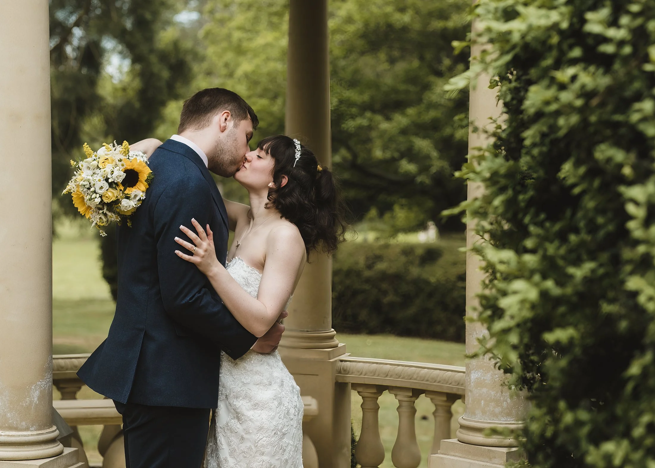 A newlywed couple shares a kiss outdoors, surrounded by greenery and classical columns, with the bride holding a bouquet of sunflowers and white flowers.