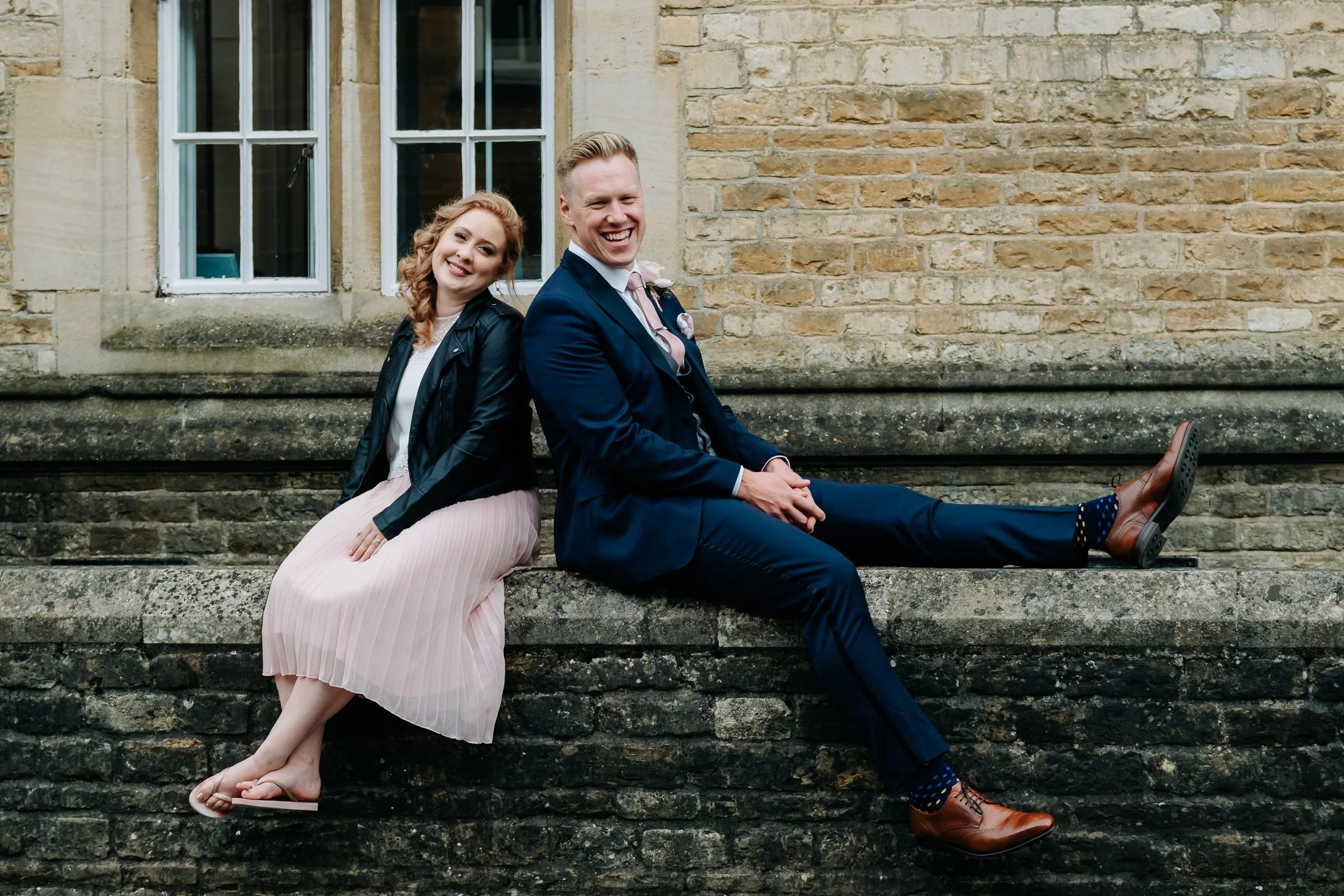 A smiling man and woman sitting back-to-back on a stone ledge in front of a brick building with large windows. The woman wears a black leather jacket and a pink pleated dress, barefoot. The man wears a navy suit, white shirt, pink tie, brown dress shoes, and patterned socks.