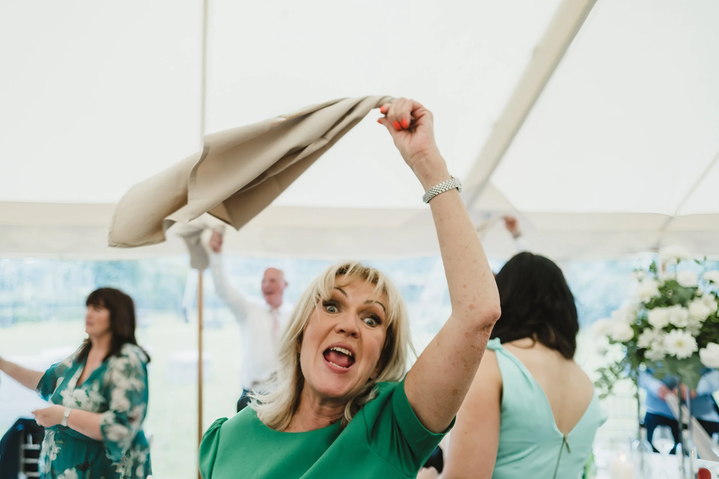 A woman in a green dress looks excited and happy, holding up a napkin or cloth during a celebration or gathering under a white tent, with other people dancing and socializing in the background.