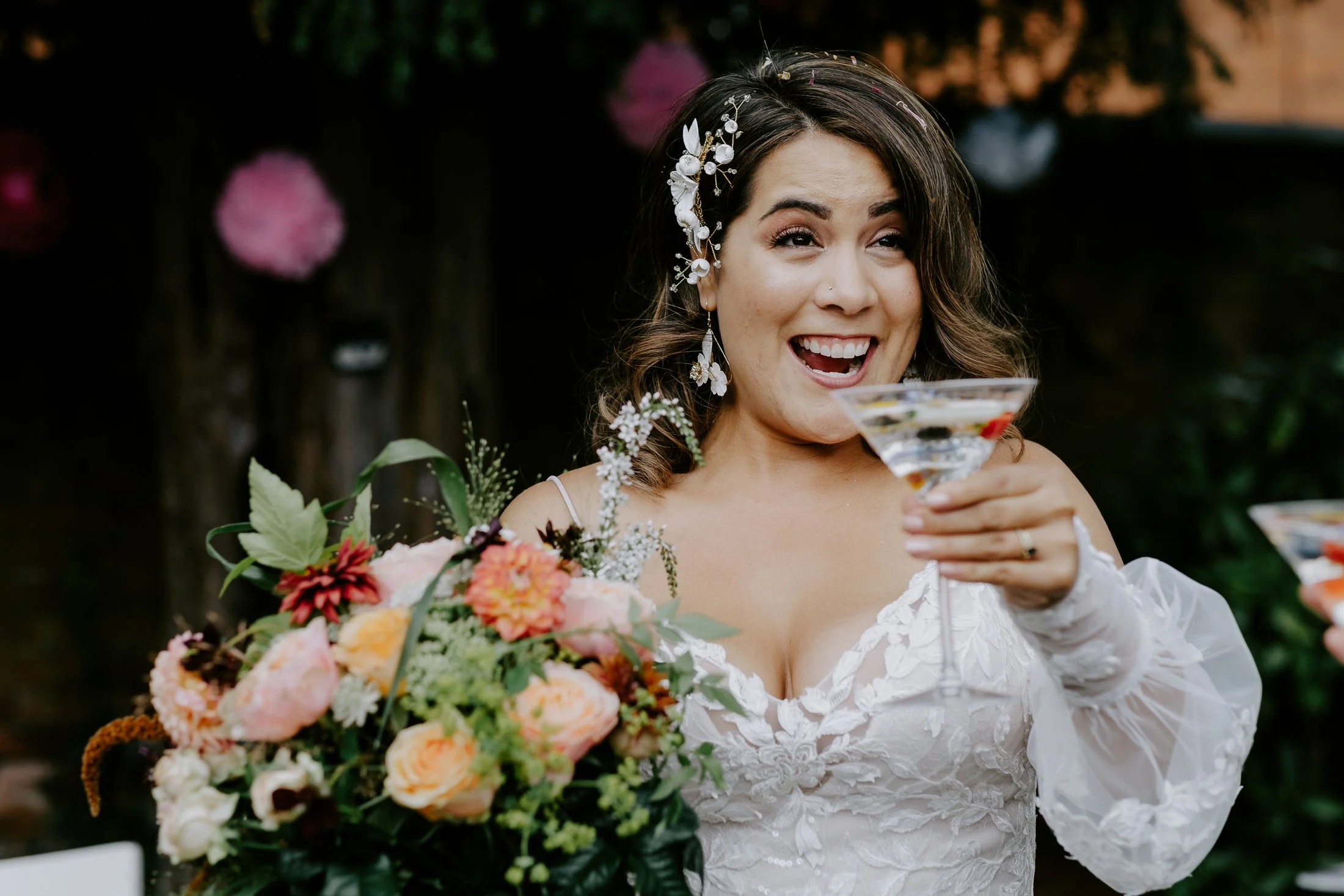Bride holding a floral bouquet and raising a cocktail glass, smiling during her wedding celebration, with a dark outdoor background.