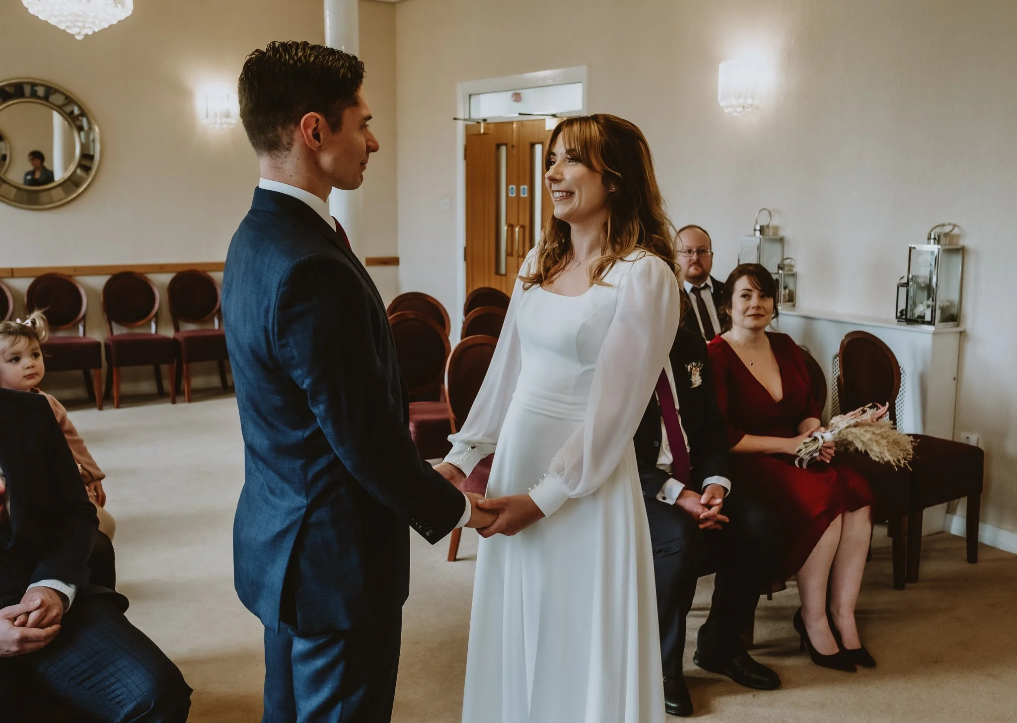 A bride and groom holding hands and looking at each other during a wedding ceremony in a warmly lit room with guests seated behind them.