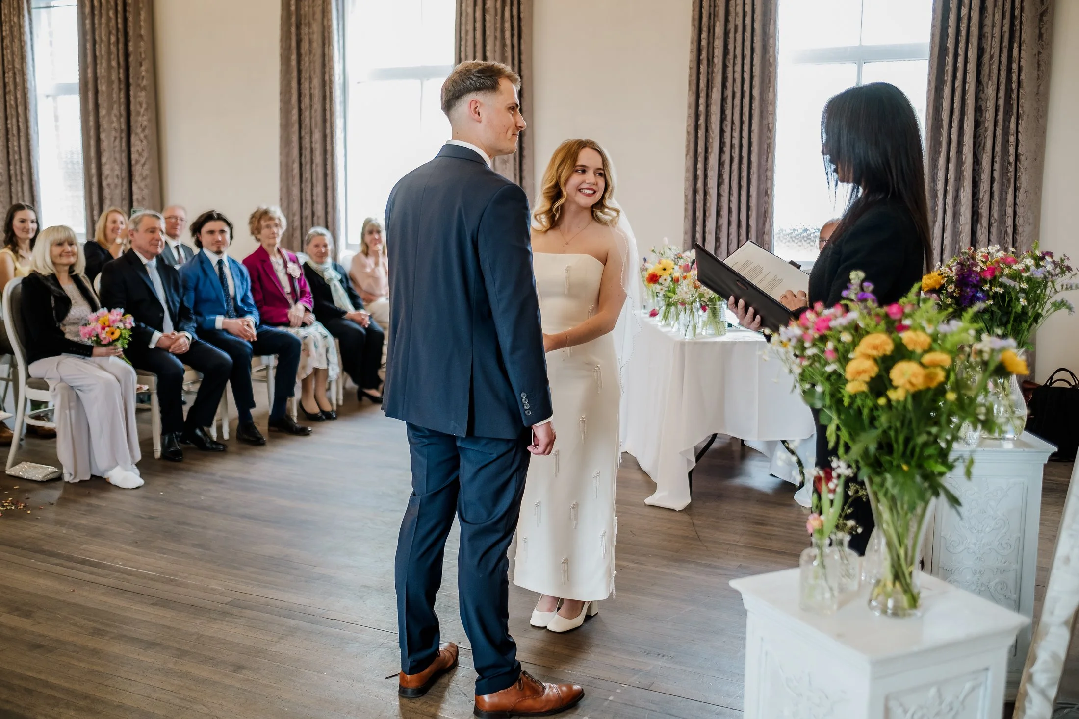 A wedding ceremony with a couple standing before an officiant, with guests seated and paying attention in the background, indoors with floral decorations.