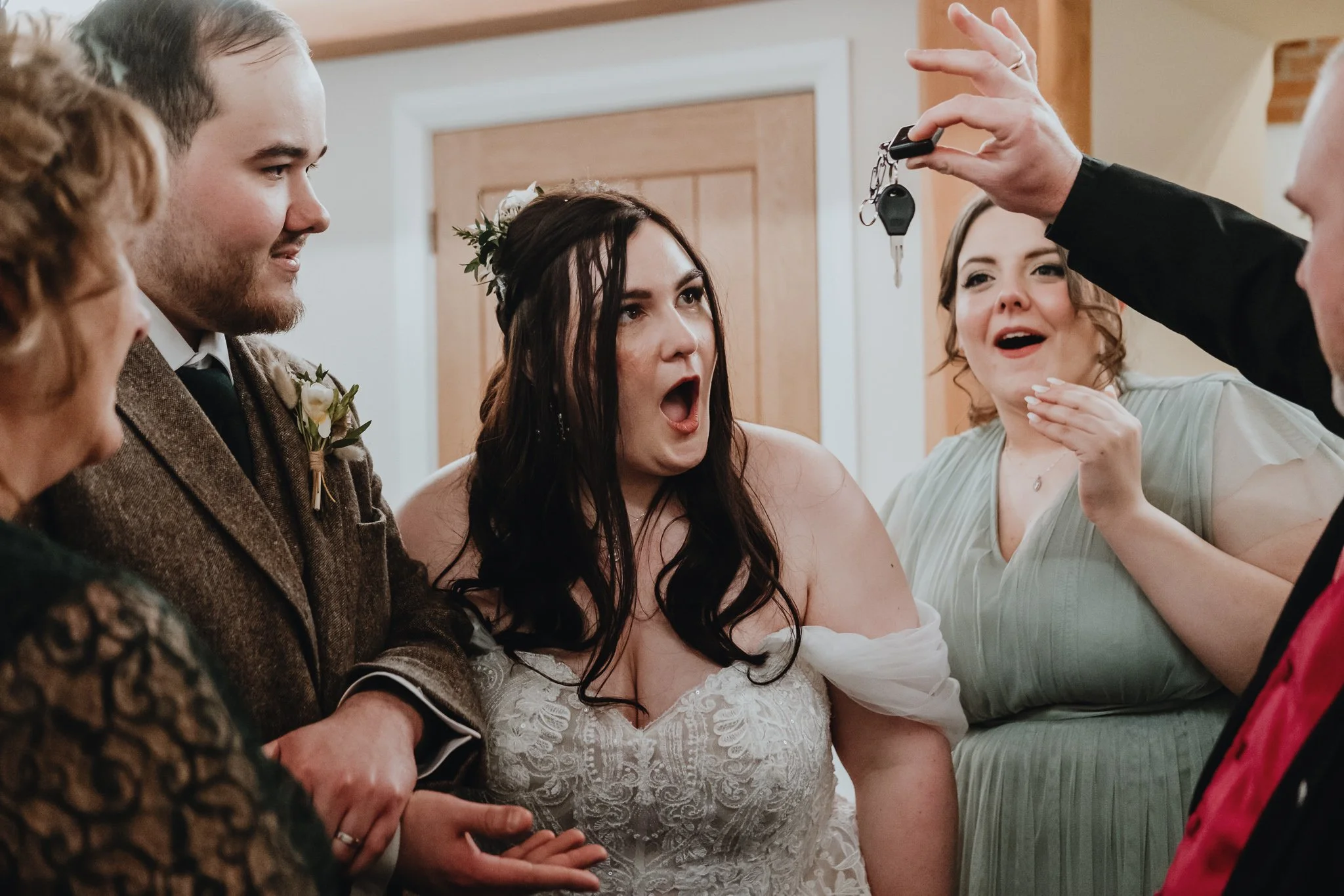 A bride with dark hair and a floral headpiece is surprised while holding hands with a groom during a gift exchange at her wedding. A man is holding out a set of keys, and a woman in a light green dress is smiling.