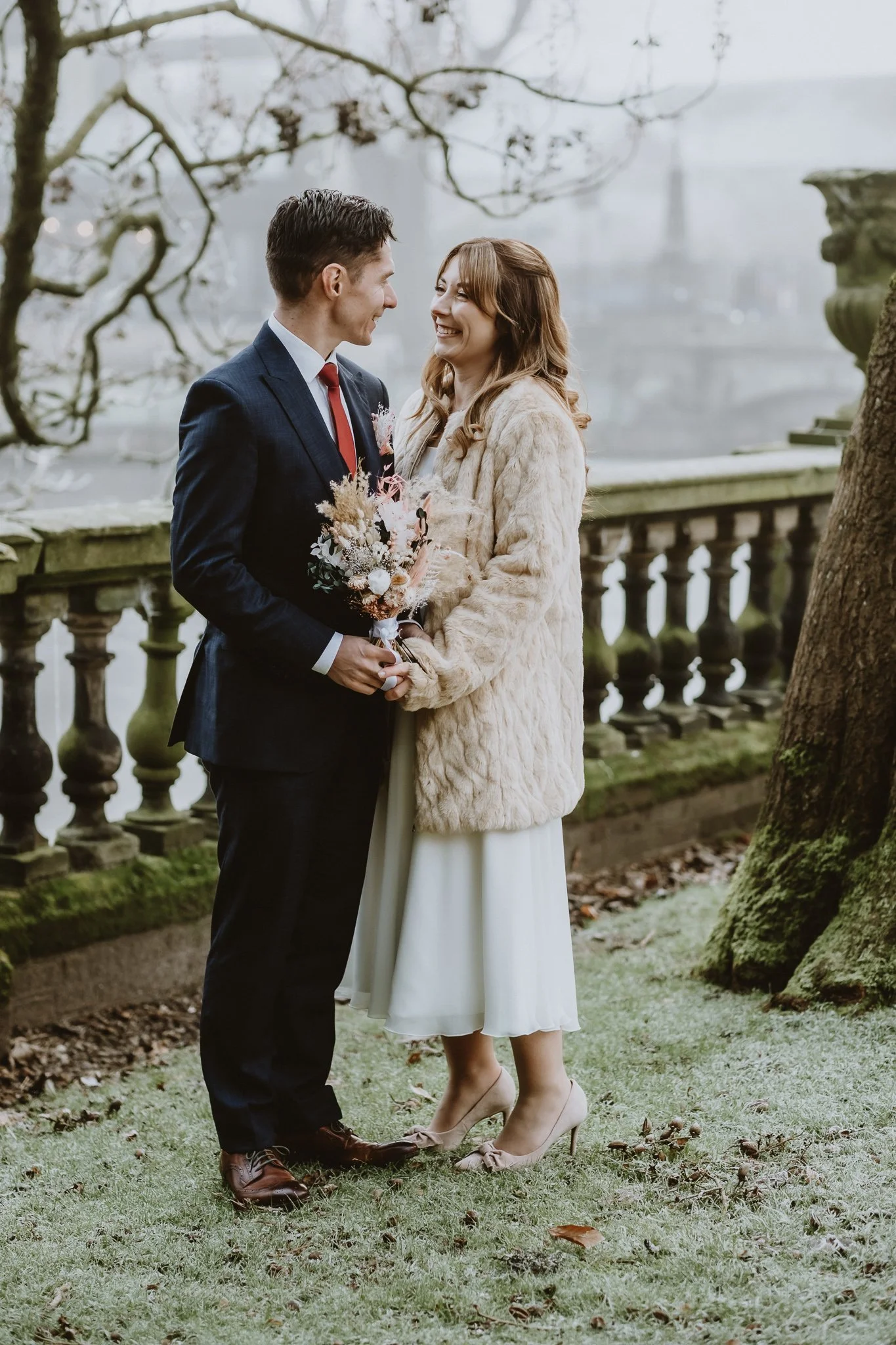 A couple dressed in wedding attire standing outdoors on a grassy area, holding a bouquet of flowers, and smiling at each other, with a stone railing and trees in the background.