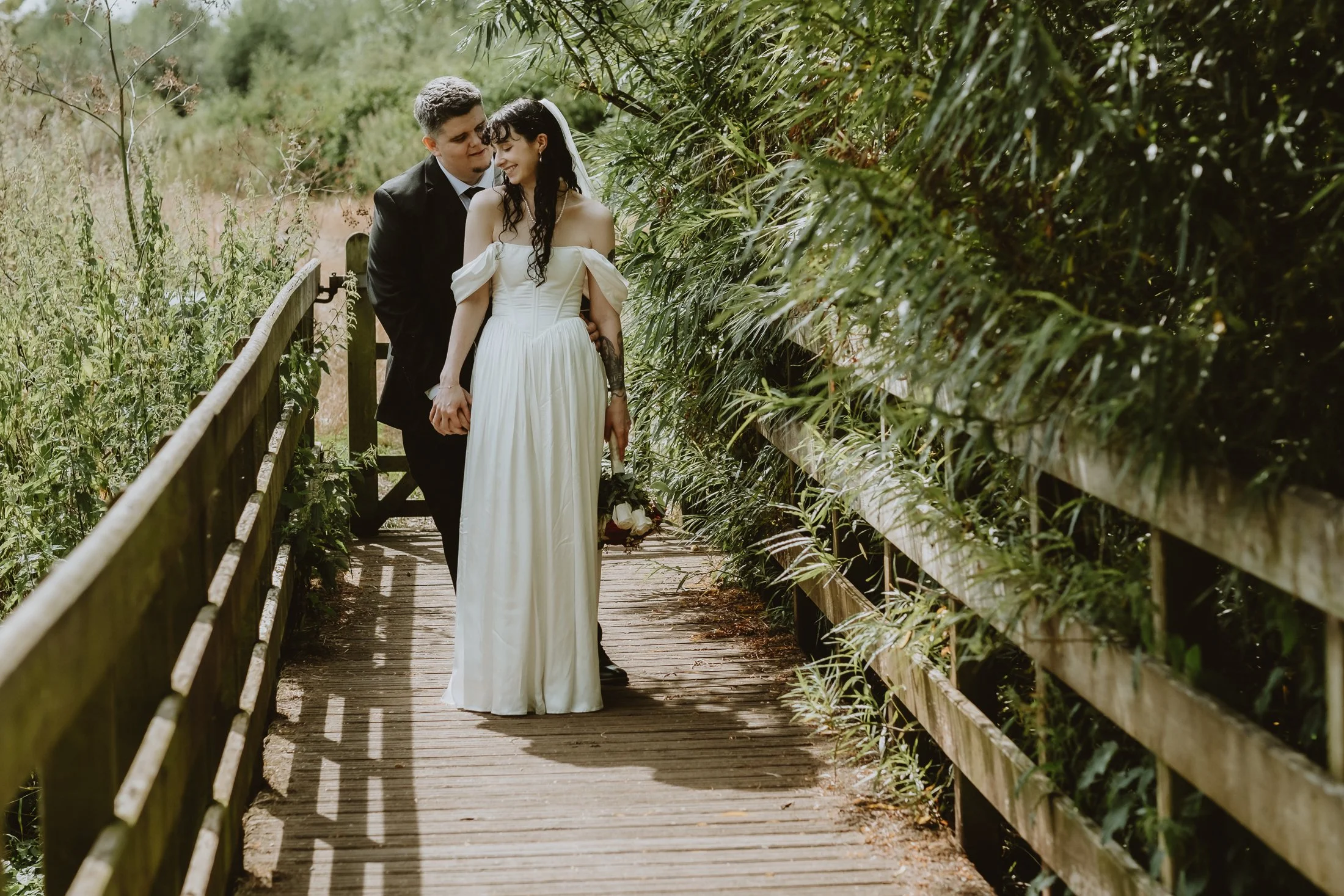A newlywed couple walking hand-in-hand on a wooden bridge surrounded by greenery, with the bride in a white dress holding a bouquet and the groom in a black suit.