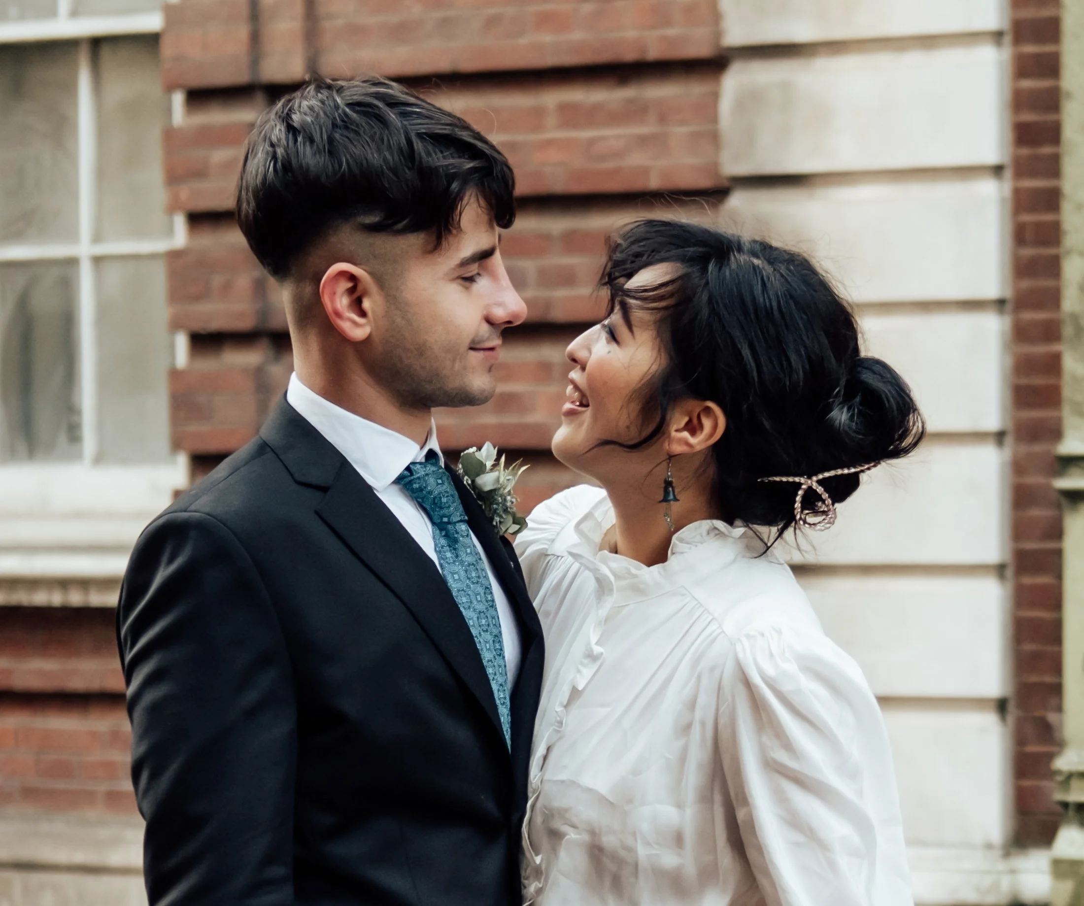 A young man in a black suit and a woman in a white blouse are facing each other, smiling, in front of a brick and stone building.