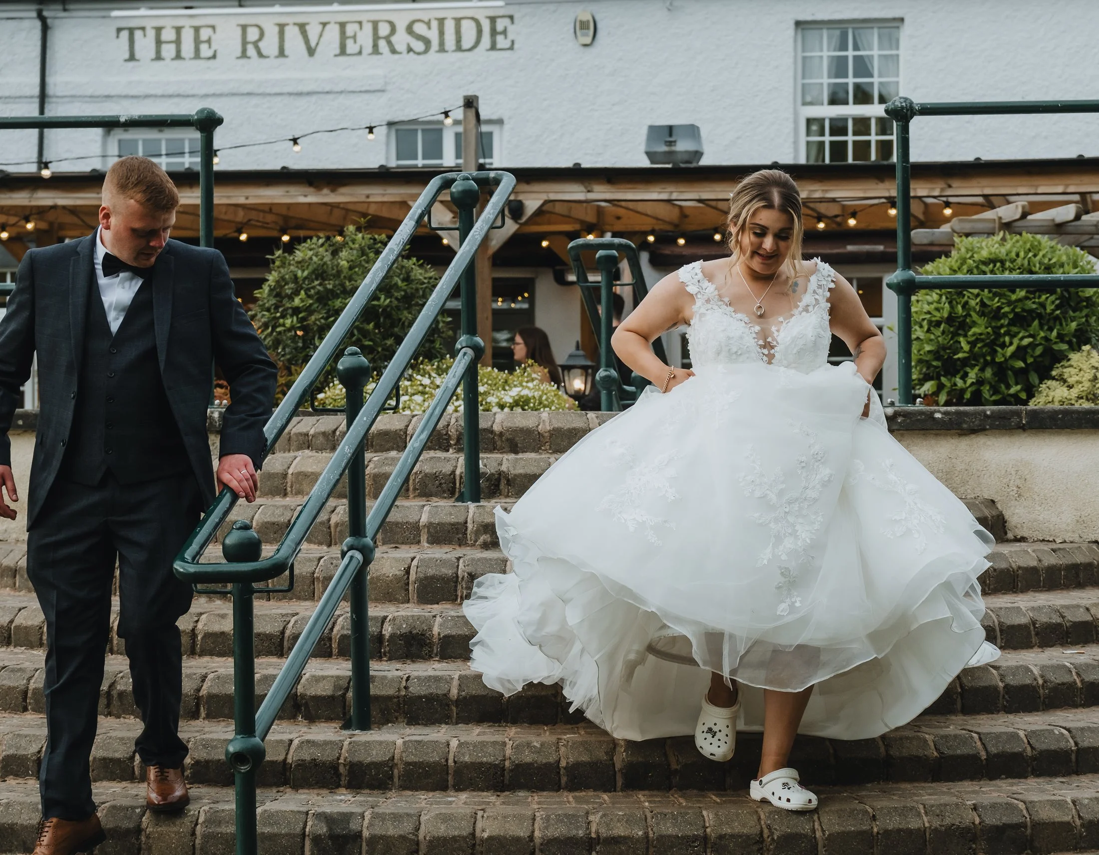 A bride in a white wedding dress with lace details, wearing white Crocs, is descending stone stairs, assisted by a groom in a dark suit. The setting is outside a building called The Riverside, with string lights and greenery in the background.