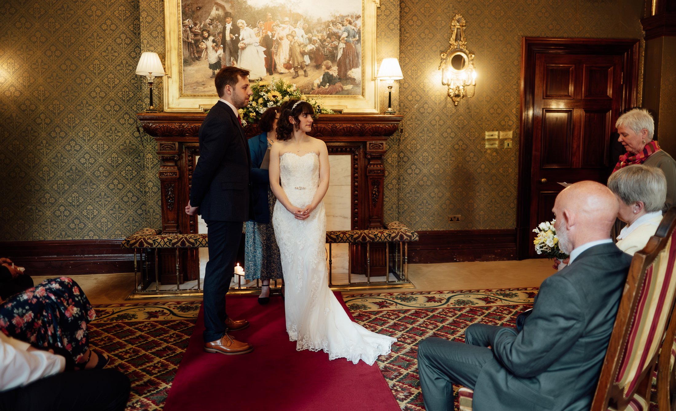 A wedding ceremony taking place indoors with a bride and groom standing before an audience, facing each other. The bride is in a white lace wedding dress, and the groom is in a dark suit. An officiant and guests are present, with some seated and one 
