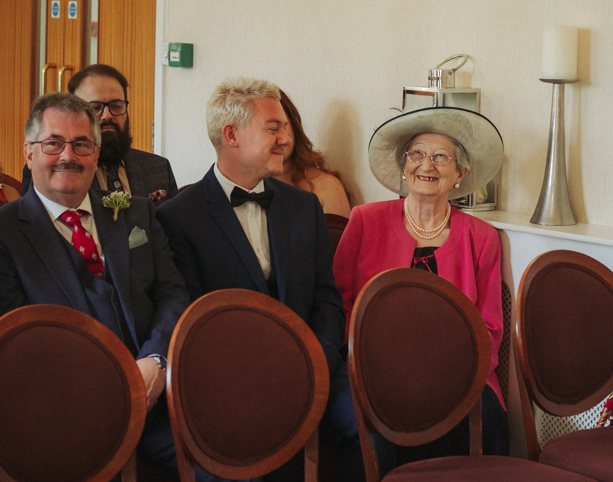Older woman wearing a large white hat, pink jacket, pearl necklace, and glasses smiling at a formal event next to a young man in a tuxedo, with other guests seated nearby.