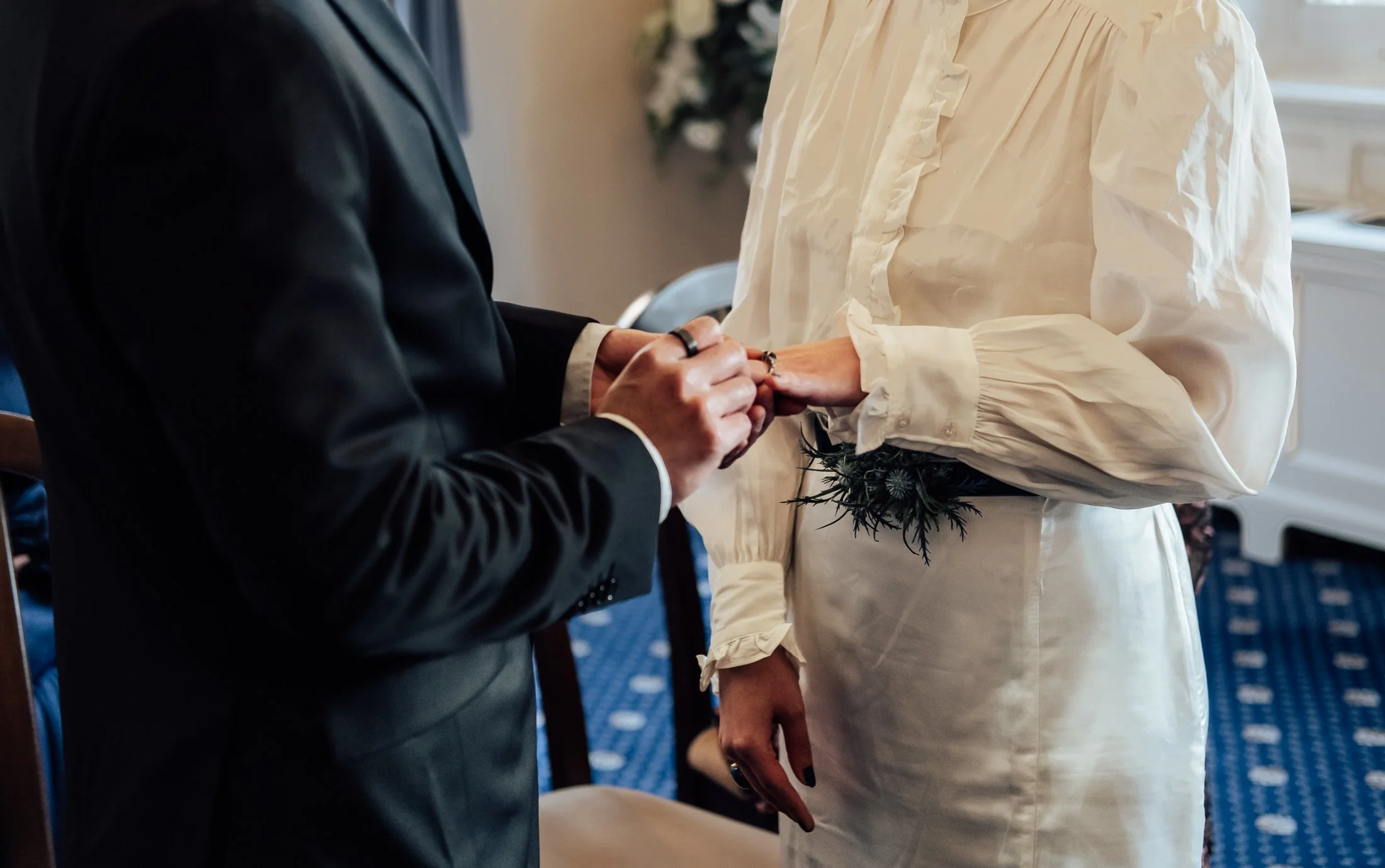 A couple holding hands during their wedding ceremony, with the bride wearing a vintage-style dress with long sleeves and the groom in a dark suit, with floral decorations visible in the background.