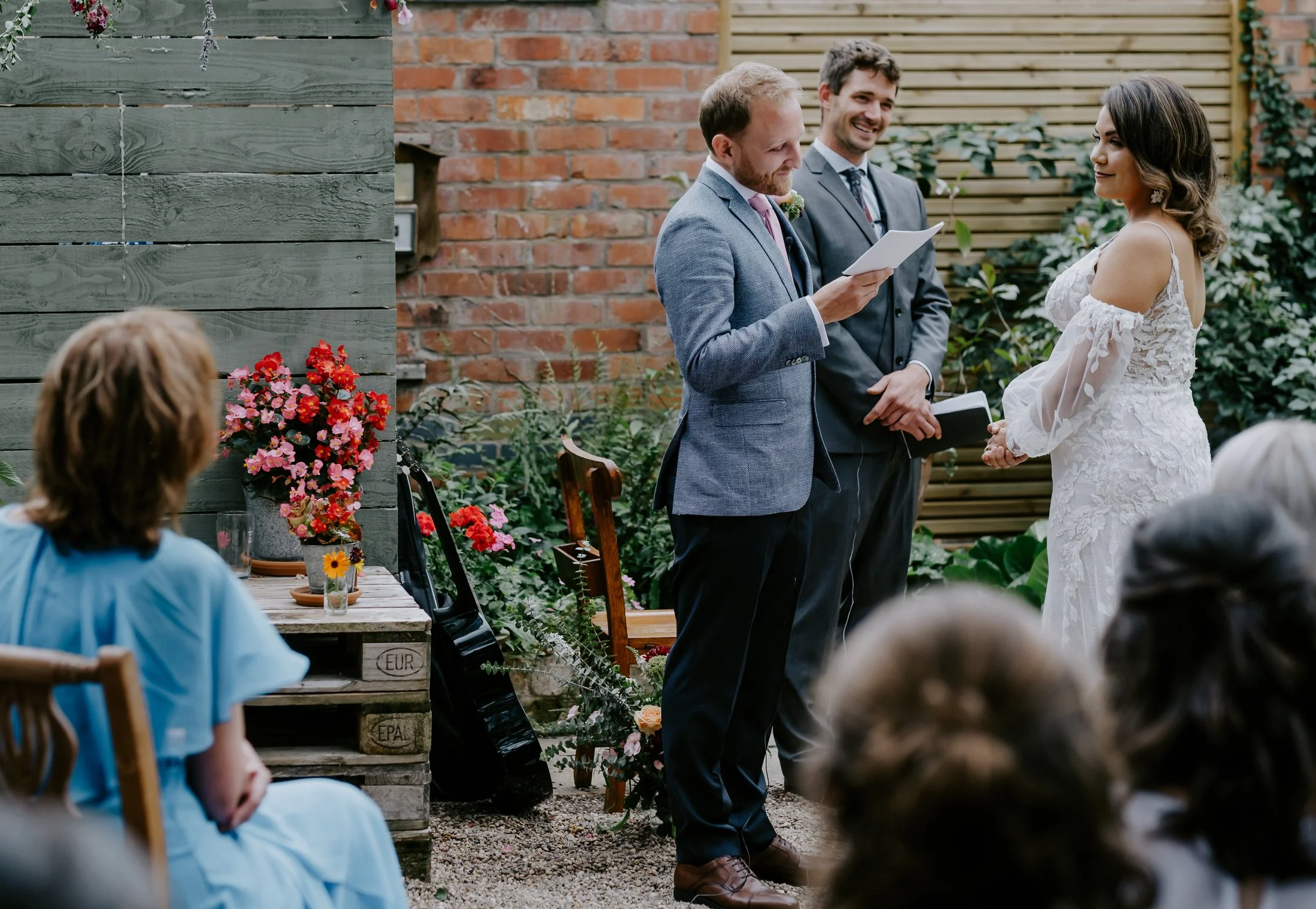 A couple getting married outdoors in front of friends, with a man reading vows or a speech, and a woman in a white wedding dress standing across from him. The setting is decorated with flowers and greenery, with a brick and wooden fence background.