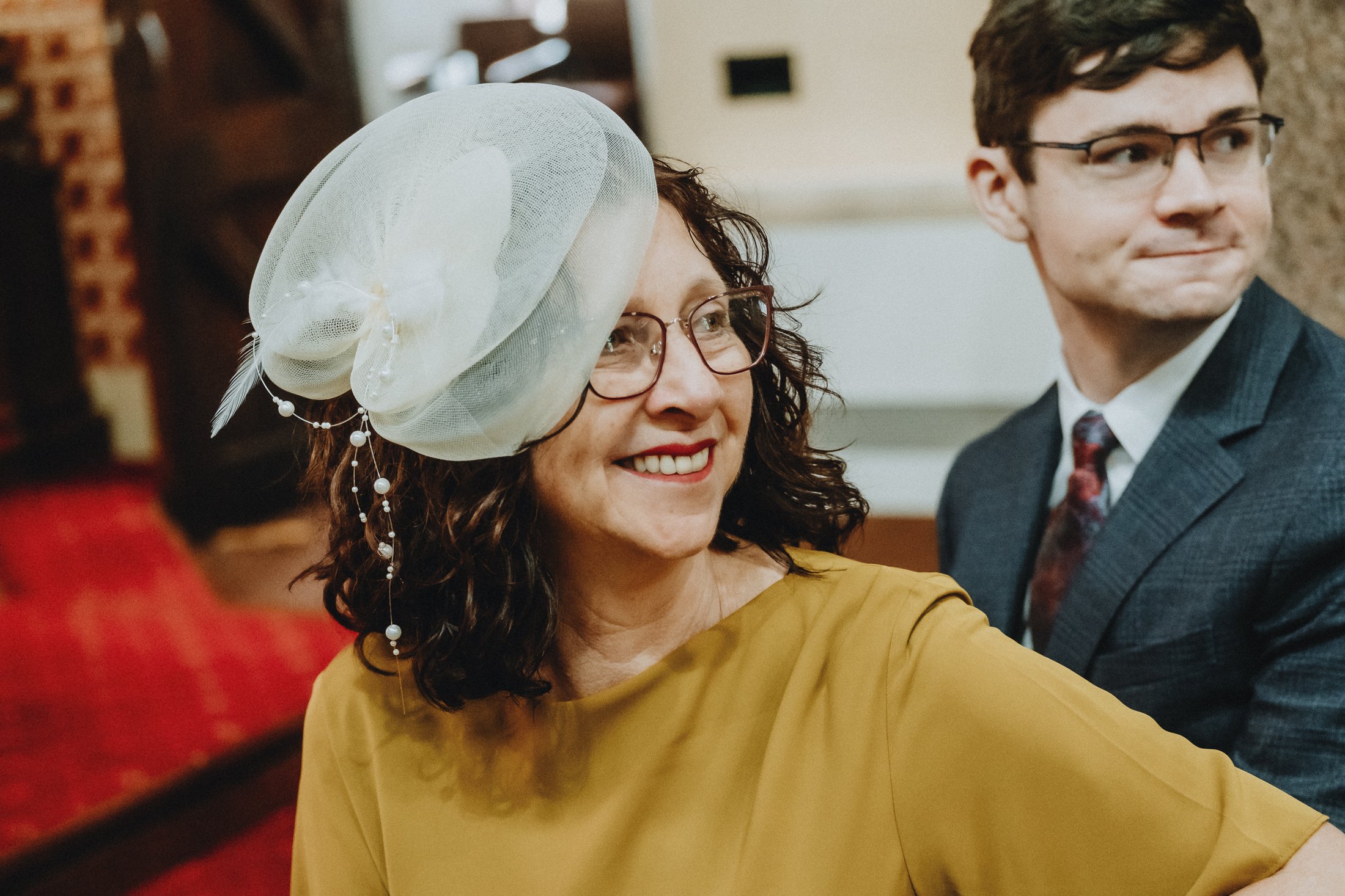 A woman with curly hair, glasses, and a yellow dress, smiling, wearing a large decorative hat with white netting, feathers, and pearl embellishments, sitting next to a man in a suit in an indoor setting.