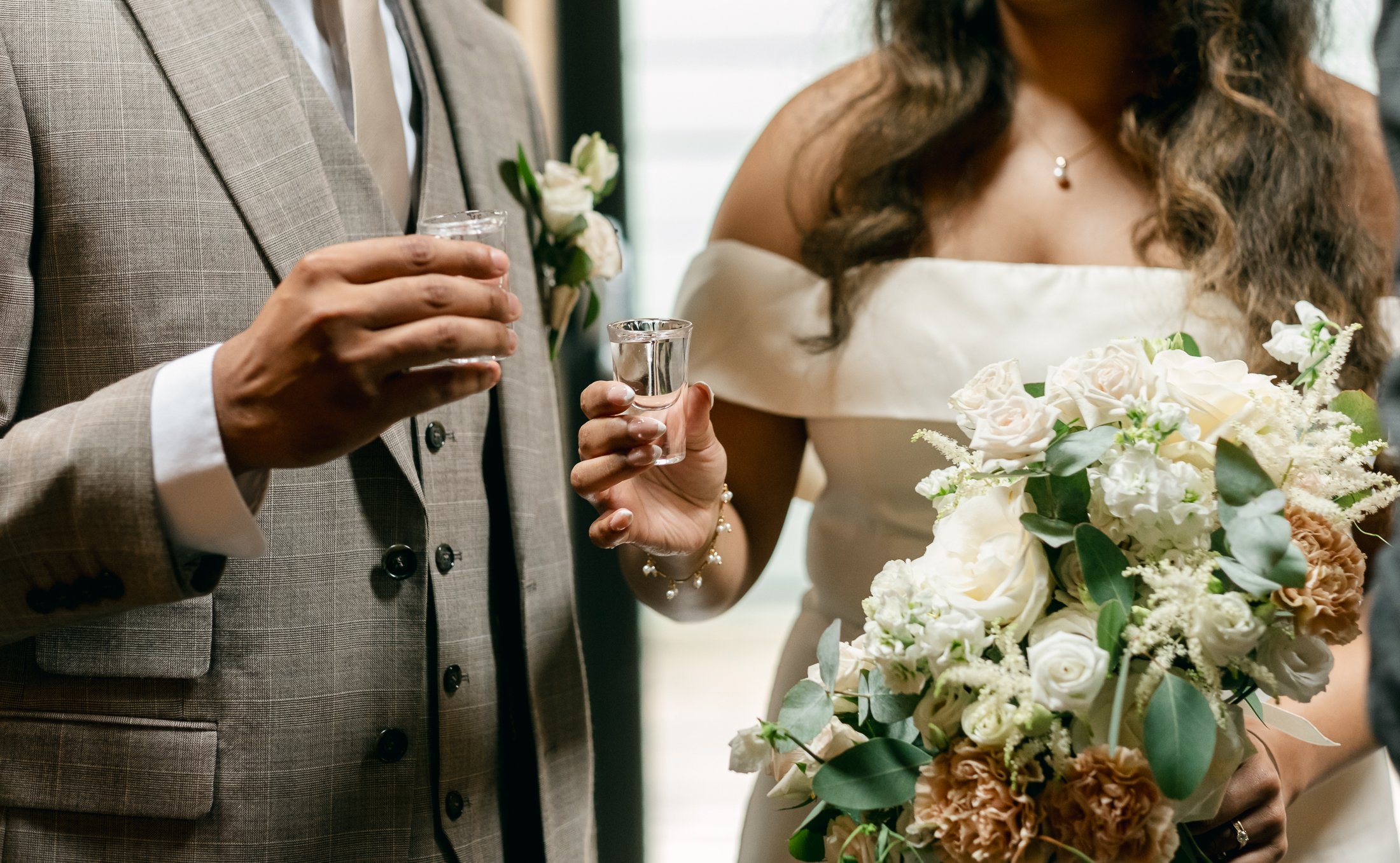 Close-up of a bride and groom holding shot glasses during a wedding celebration, with focus on their hands, floral wedding bouquet, and formal attire.