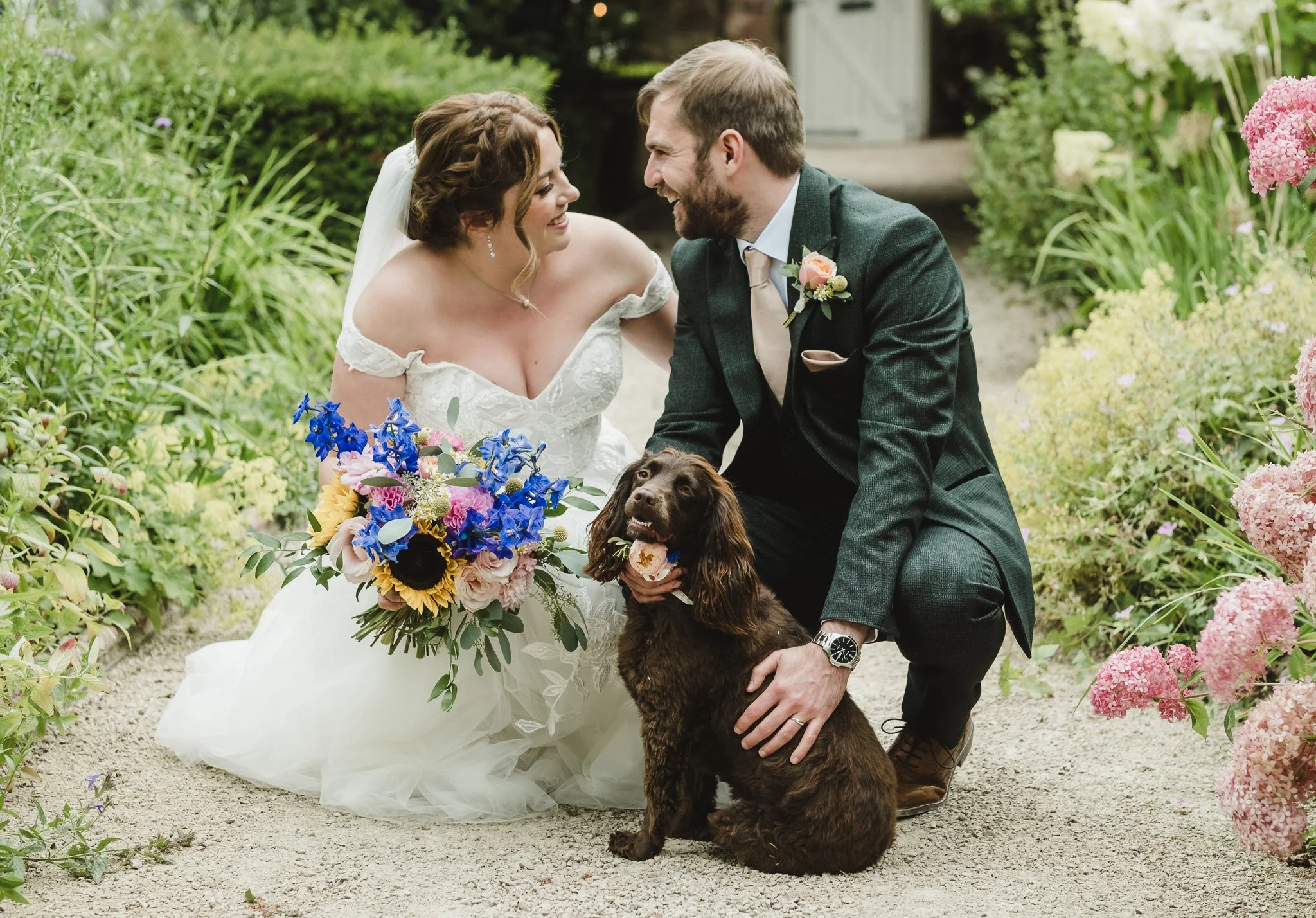 A bride and groom kneeling on a garden path, smiling at each other, with a brown spaniel dog between them, surrounded by colorful flowers.