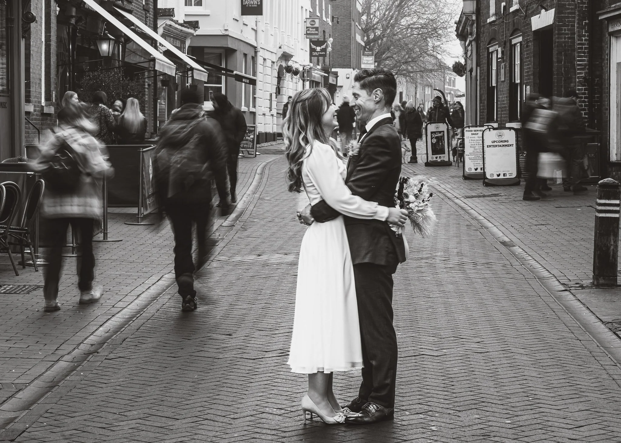 A couple dressed in wedding attire sharing a romantic moment on a city street, with people walking past in motion blur.
