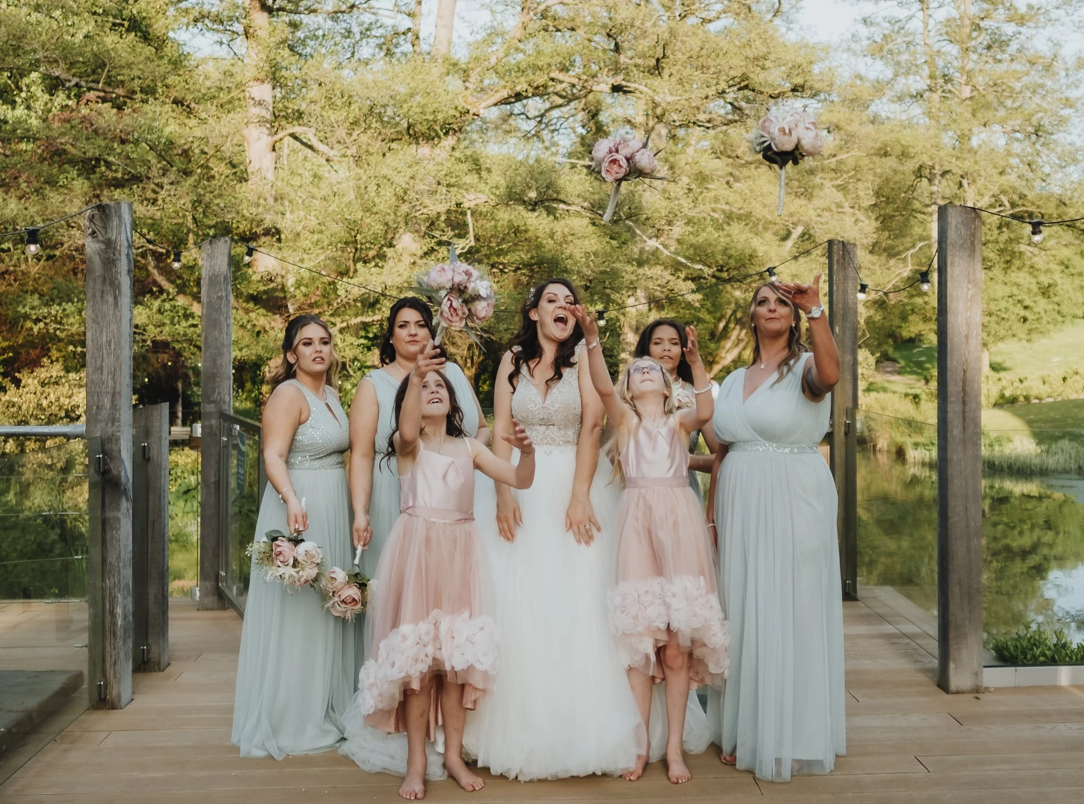 A bride and her bridesmaids and flower girls standing outdoors on a wooden deck, dressed in pastel-colored dresses, with some holding flower bouquets, tossing flower petals or flowers into the air during a wedding celebration.