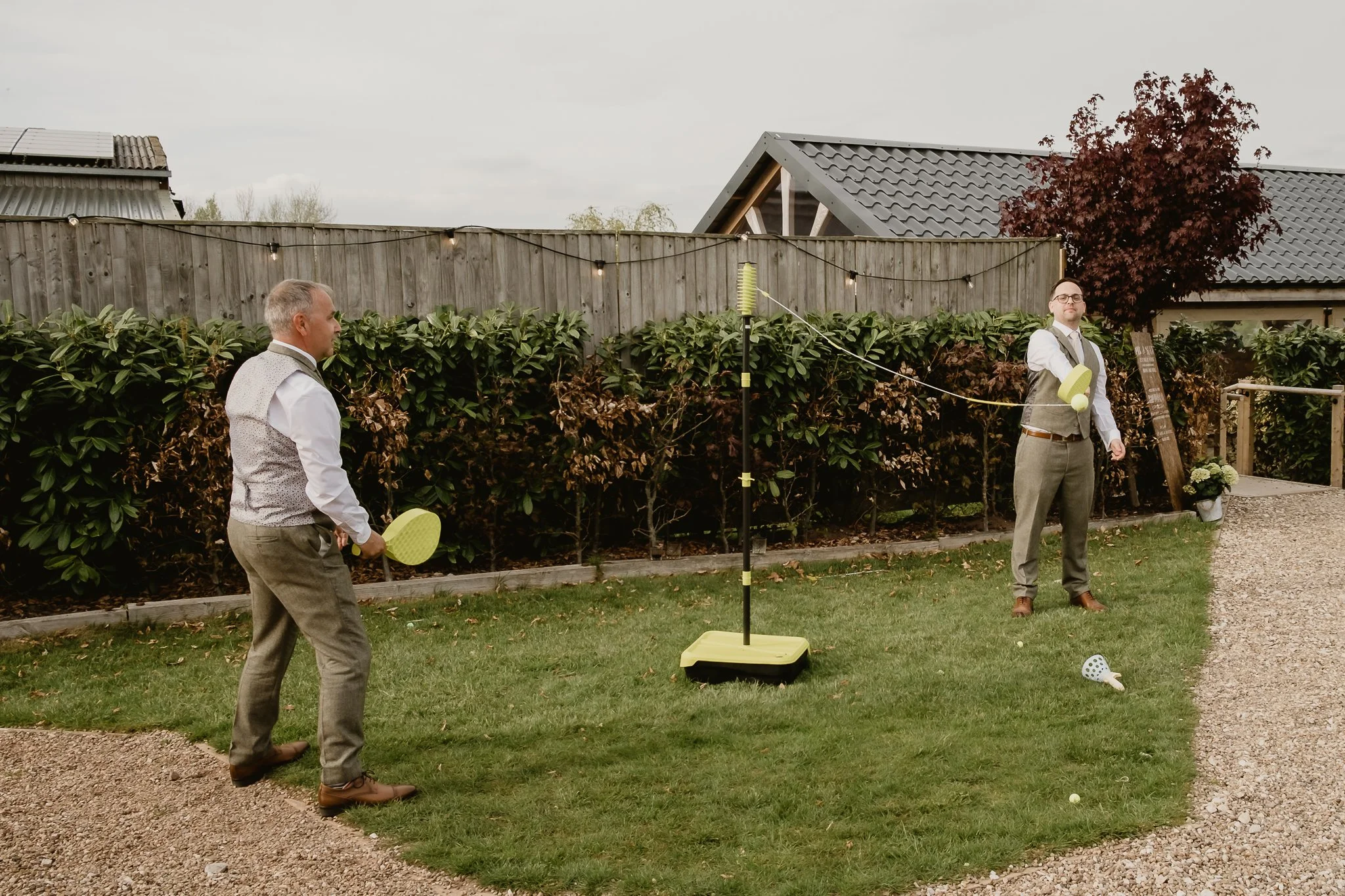 Two men in formal attire playing badminton outdoors in a backyard. One man is preparing to serve, while the other stands ready. The net is set up in front of a hedge, with houses and trees in the background.