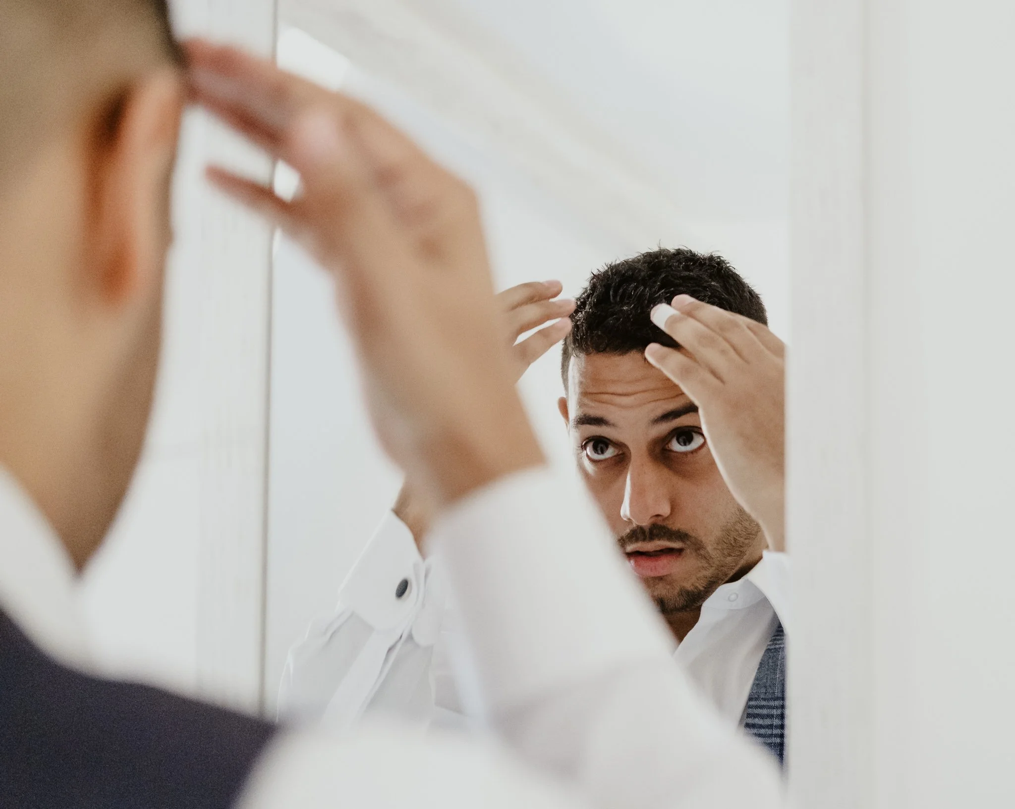 A man with dark hair and a white shirt looks into a mirror while styling his hair, with his hands on his head.