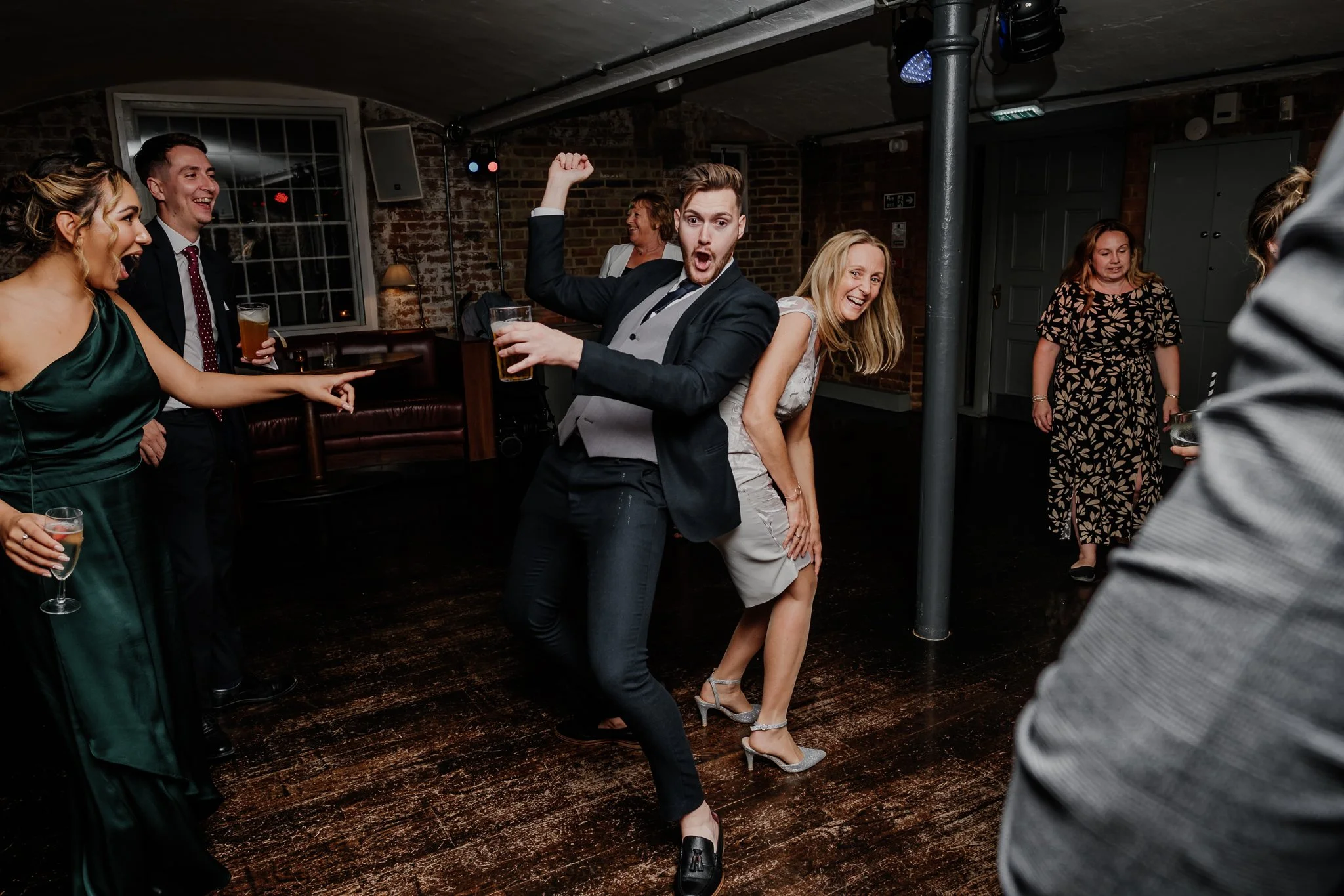 People dancing and having fun at a party in a dimly lit room with brick walls, some holding drinks