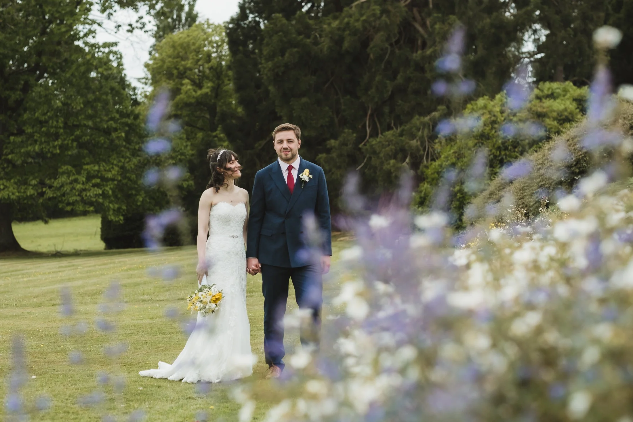 A bride and groom holding hands on a grassy field, with trees in the background, surrounded by blurred purple and white flowers.