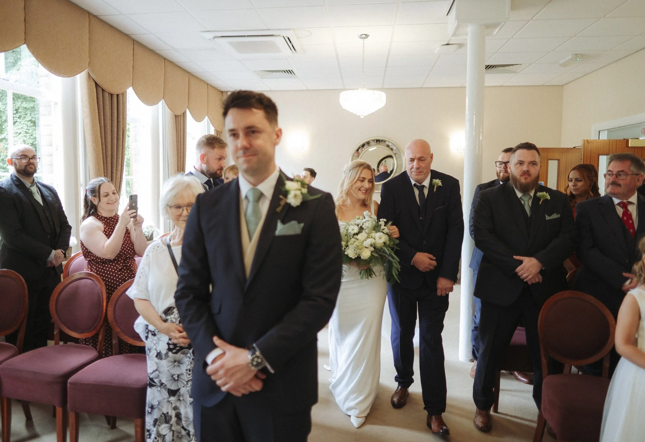 A wedding ceremony with guests seated and standing in a decorated hall. The bride is smiling and holding a bouquet of flowers, and the groom stands at the front.