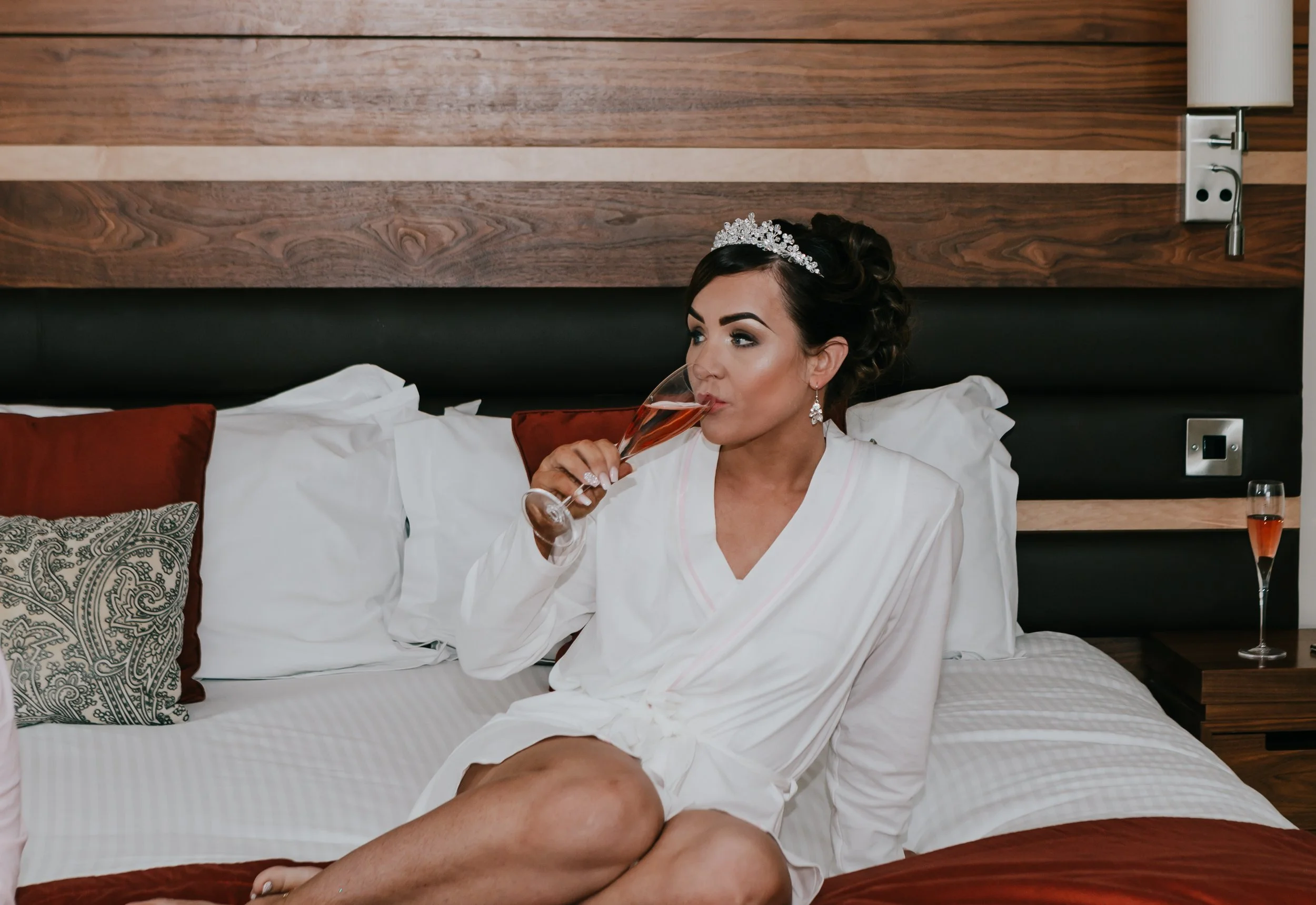 A woman wearing a white robe, a tiara, and earrings is sitting on a bed, sipping champagne from a flute in a hotel room.