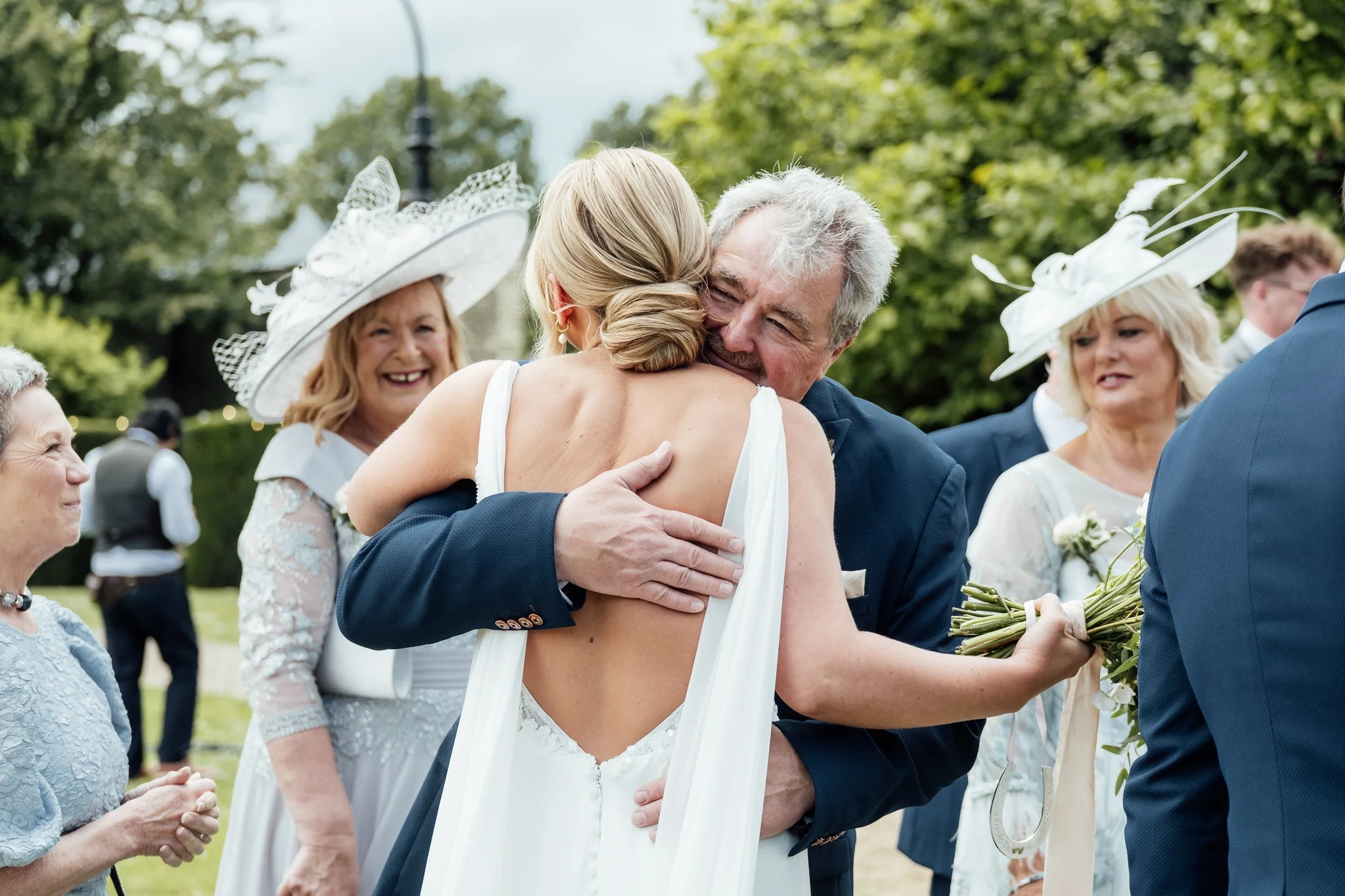 Person hugging bride at wedding ceremony outdoors with guests in the background.