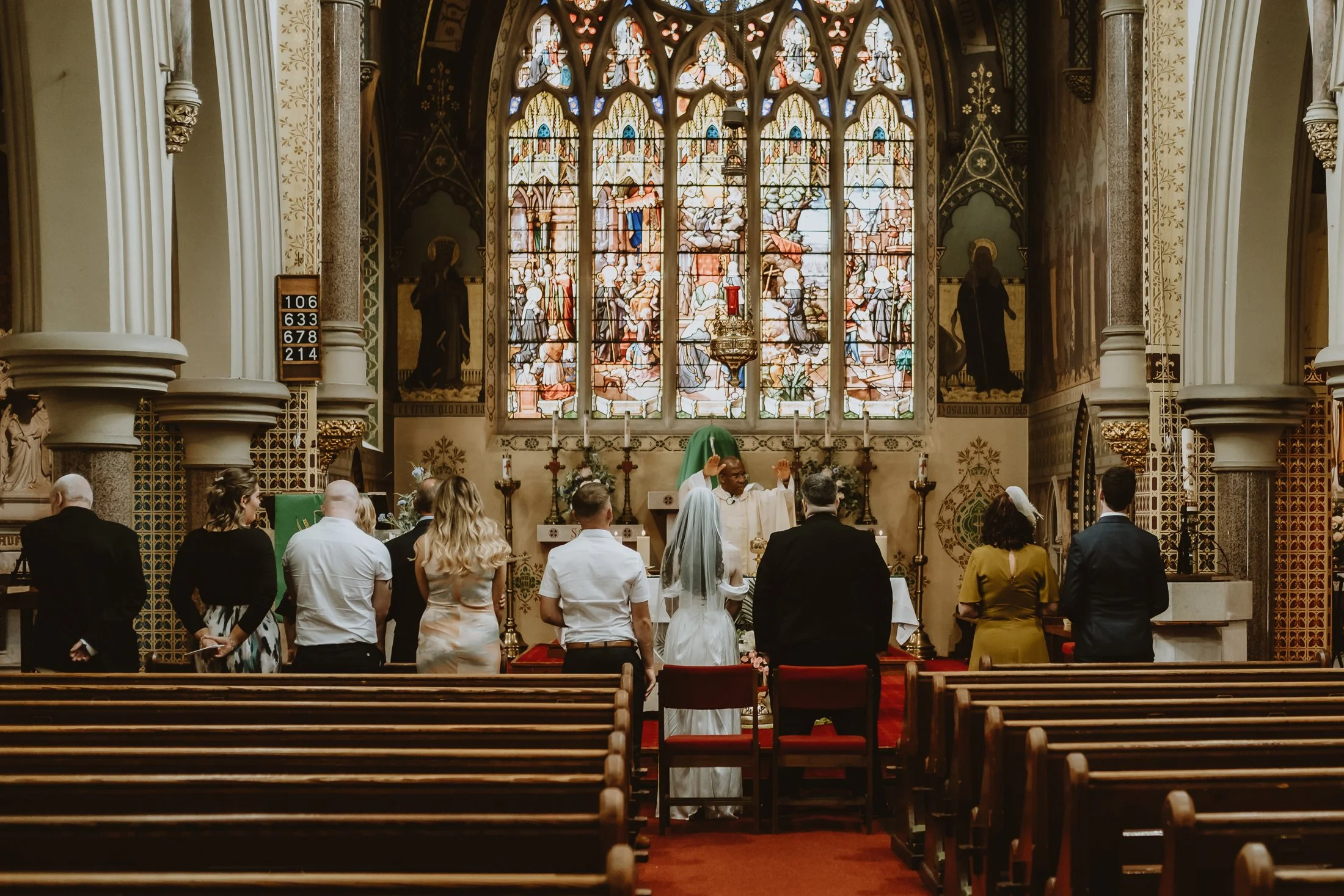 A wedding ceremony taking place inside a church with a bride and groom standing before an officiant at the altar, surrounded by guests. The church features stained glass windows and ornate decorations.