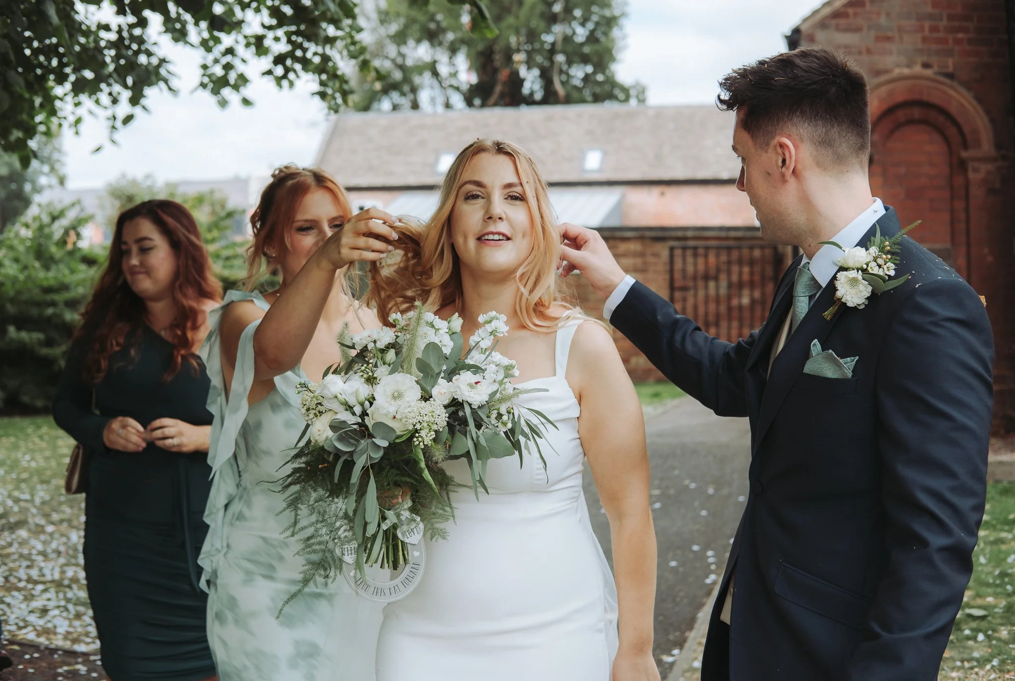 A woman in a white dress holding a bouquet of white flowers, smiling, as a man in a dark suit with a boutonnière adjusts her hair. Two women stand behind them, one with reddish hair and the other with brown hair, in an outdoor setting with trees and 