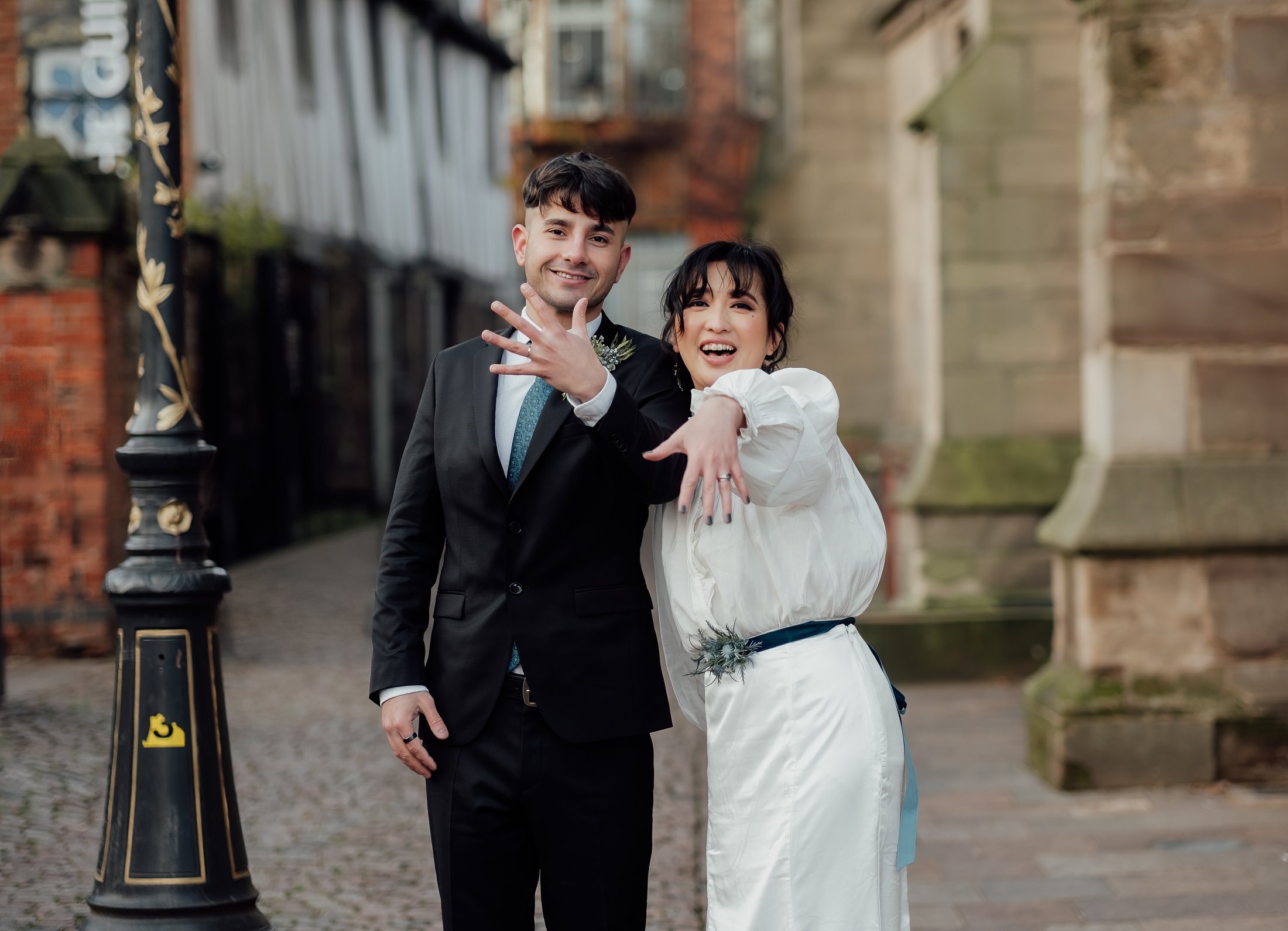A happy couple celebrating their wedding outdoors, with the groom in a black suit and the bride in a white dress, both showing off their engagement rings.