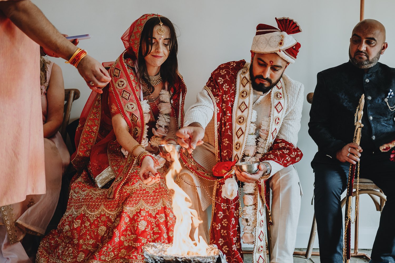 bride and groom participating in a hindu wedding ceremony, with the bride holding a brass plate with fire and the groom churning a small pot, both dressed in traditional red and white wedding attire, surrounded by seated guests.