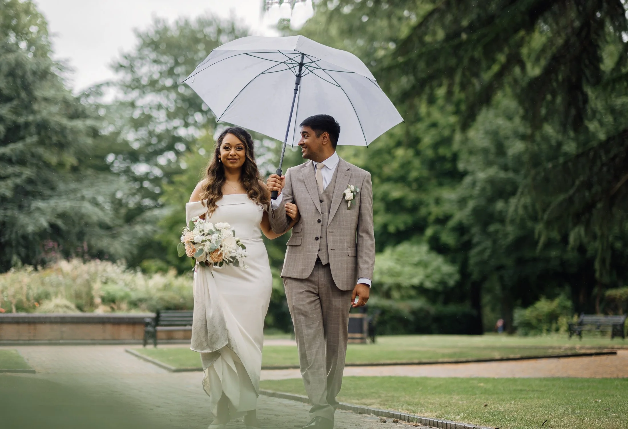 A bride and groom walking together in a park during rain, with the groom holding a white umbrella. The bride wears a white wedding dress and holds a bouquet, while the groom is in a beige suit with a boutonniere.