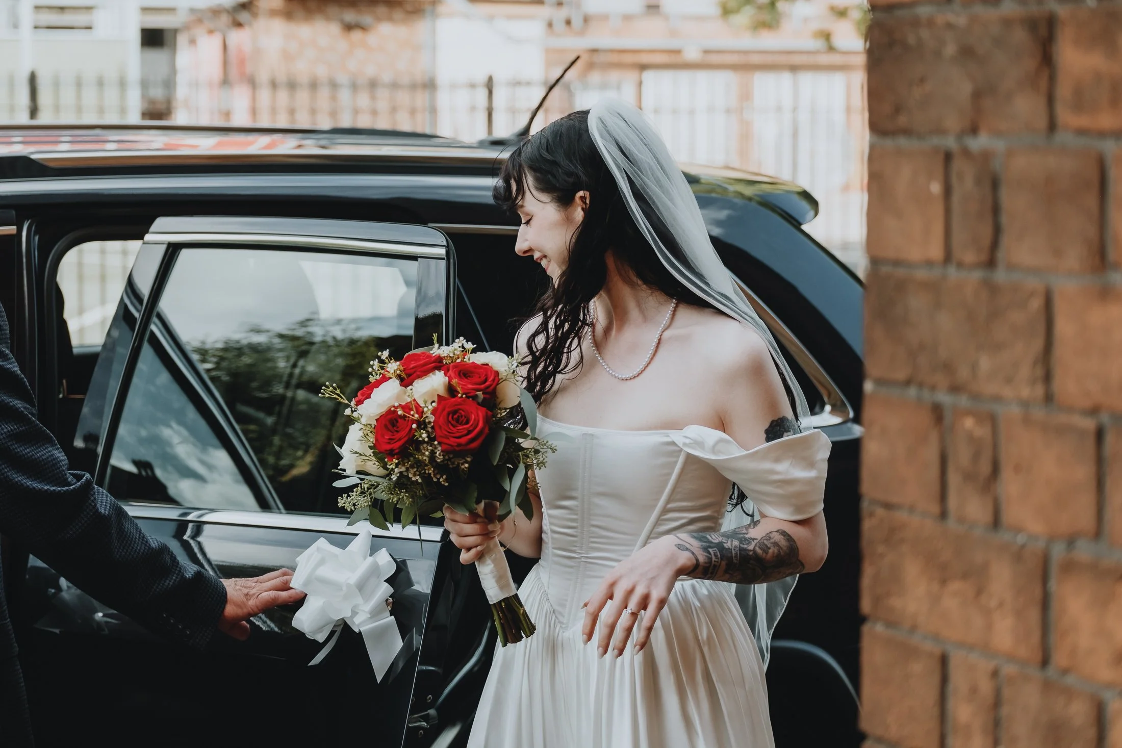 Bride in wedding dress holding a bouquet of red and white roses, smiling while exiting a black car, with a brick wall on the right side of the image.