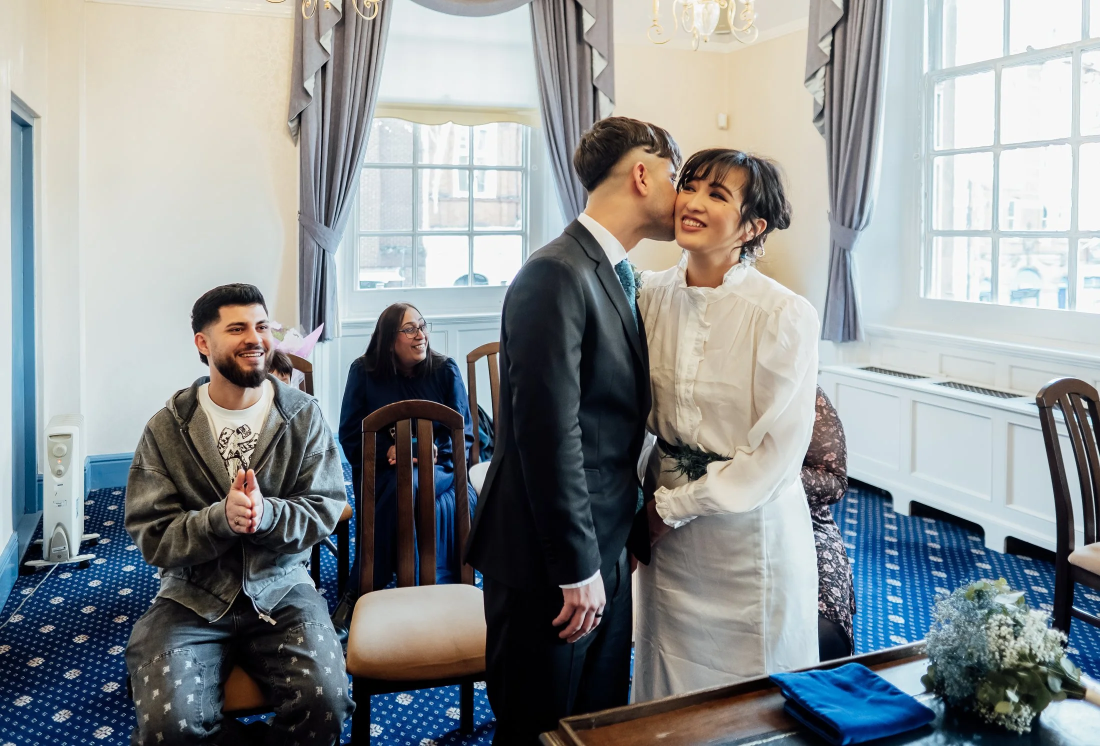 A couple is at the center of the room, with the man wearing a suit and the woman wearing a white dress, sharing a kiss on the cheek during a wedding ceremony. Two people seated in the background are smiling and looking at the couple, the man with cla