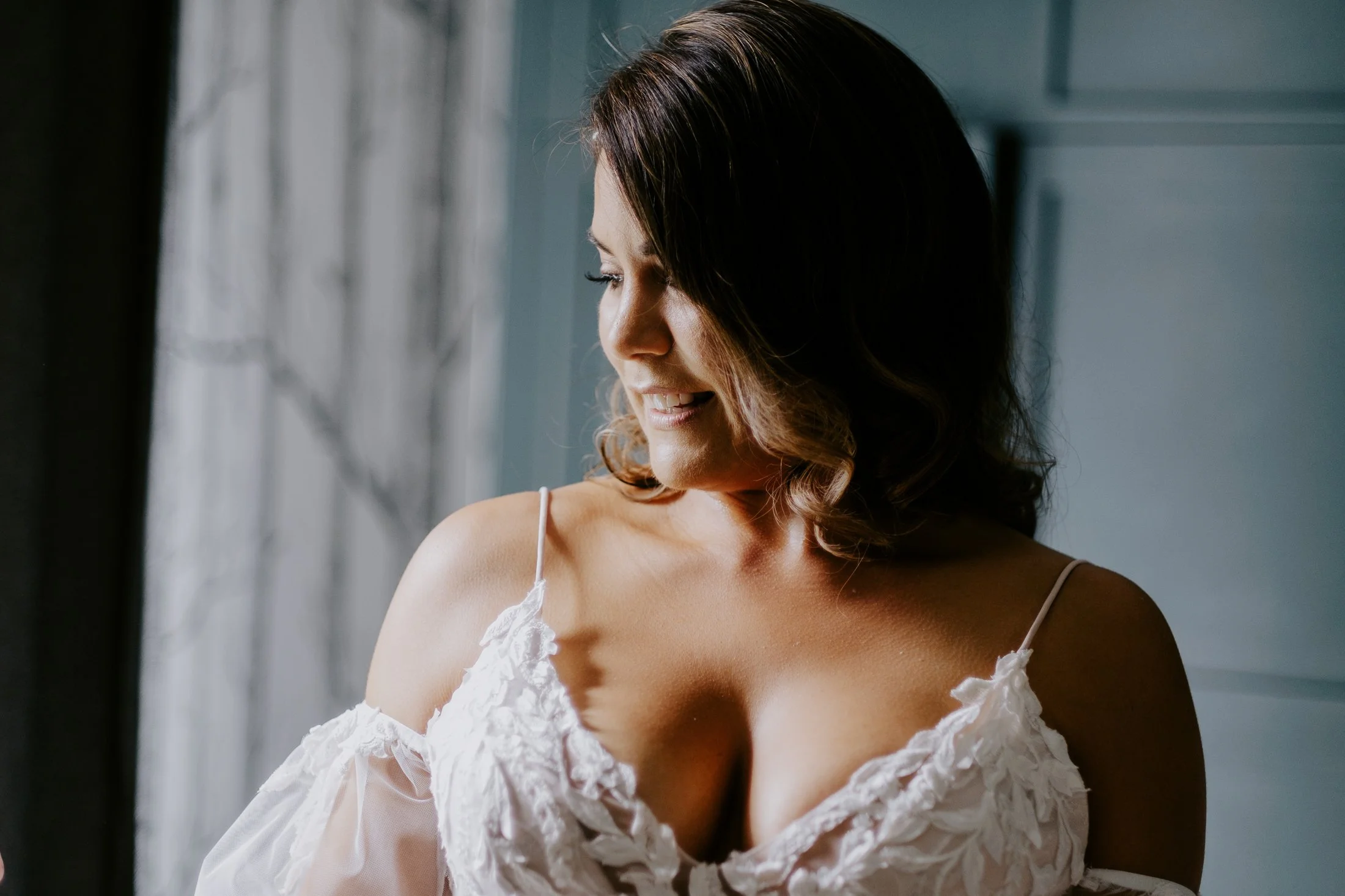 Woman with dark brown hair smiling, looking down, wearing a white off-the-shoulder dress with lace details, standing near a window with natural light.