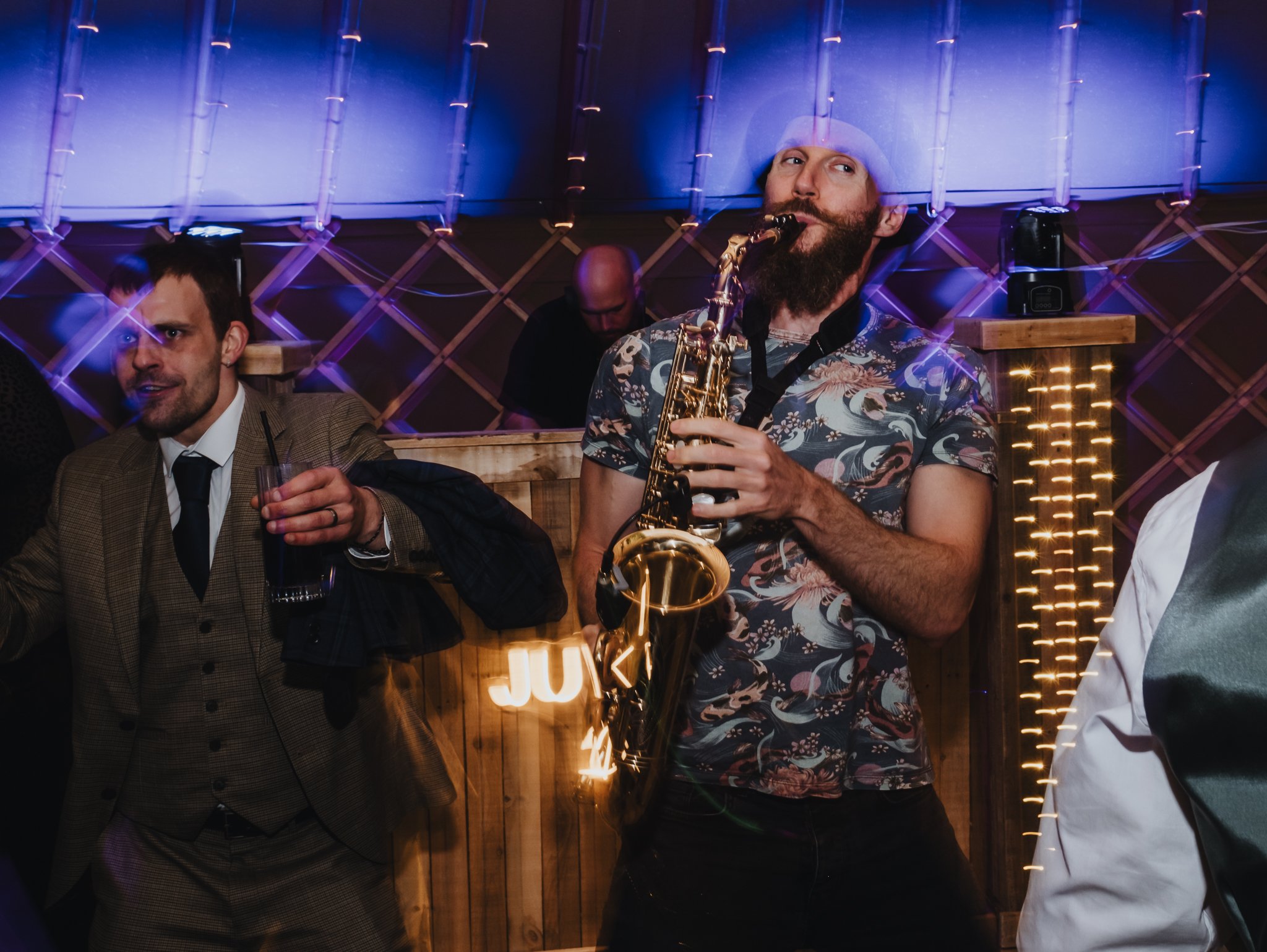A man with a beard wearing a floral shirt playing the saxophone at a lively party or bar, with other people around and colorful lighting.