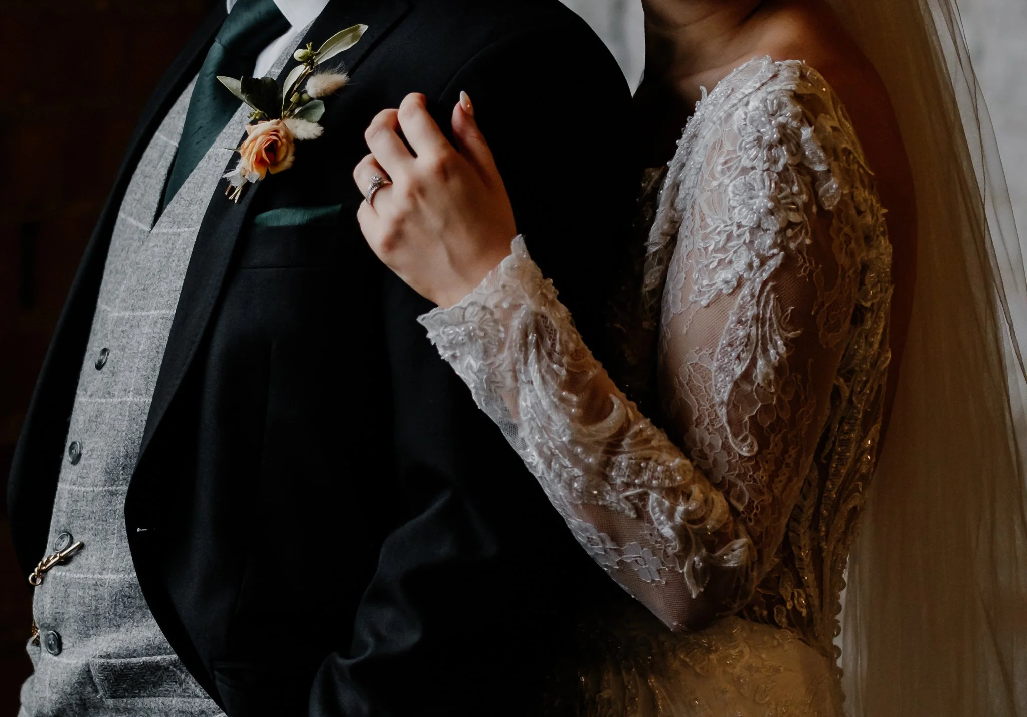 Close-up of a couple at a wedding, with the bride gently holding the groom's shoulder, wearing a lace wedding dress, and the groom in a tuxedo with a boutonniere.