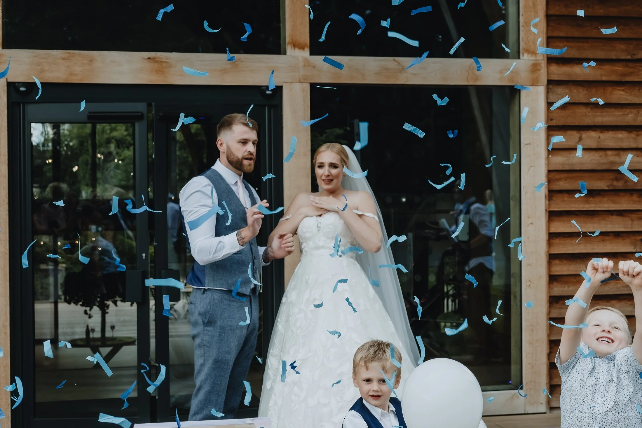 A newlywed couple celebrates with children and blue confetti outside a building with glass doors and windows.