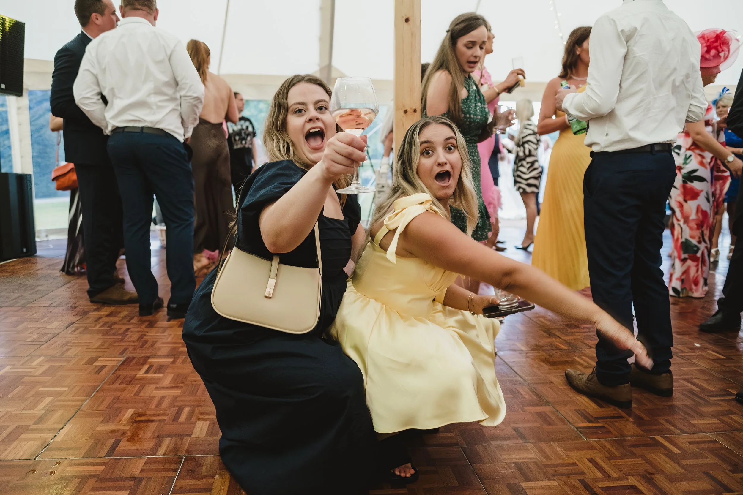 Two women sitting on the dance floor at a lively party or wedding reception, with one holding a glass of wine and both appearing surprised or excited.