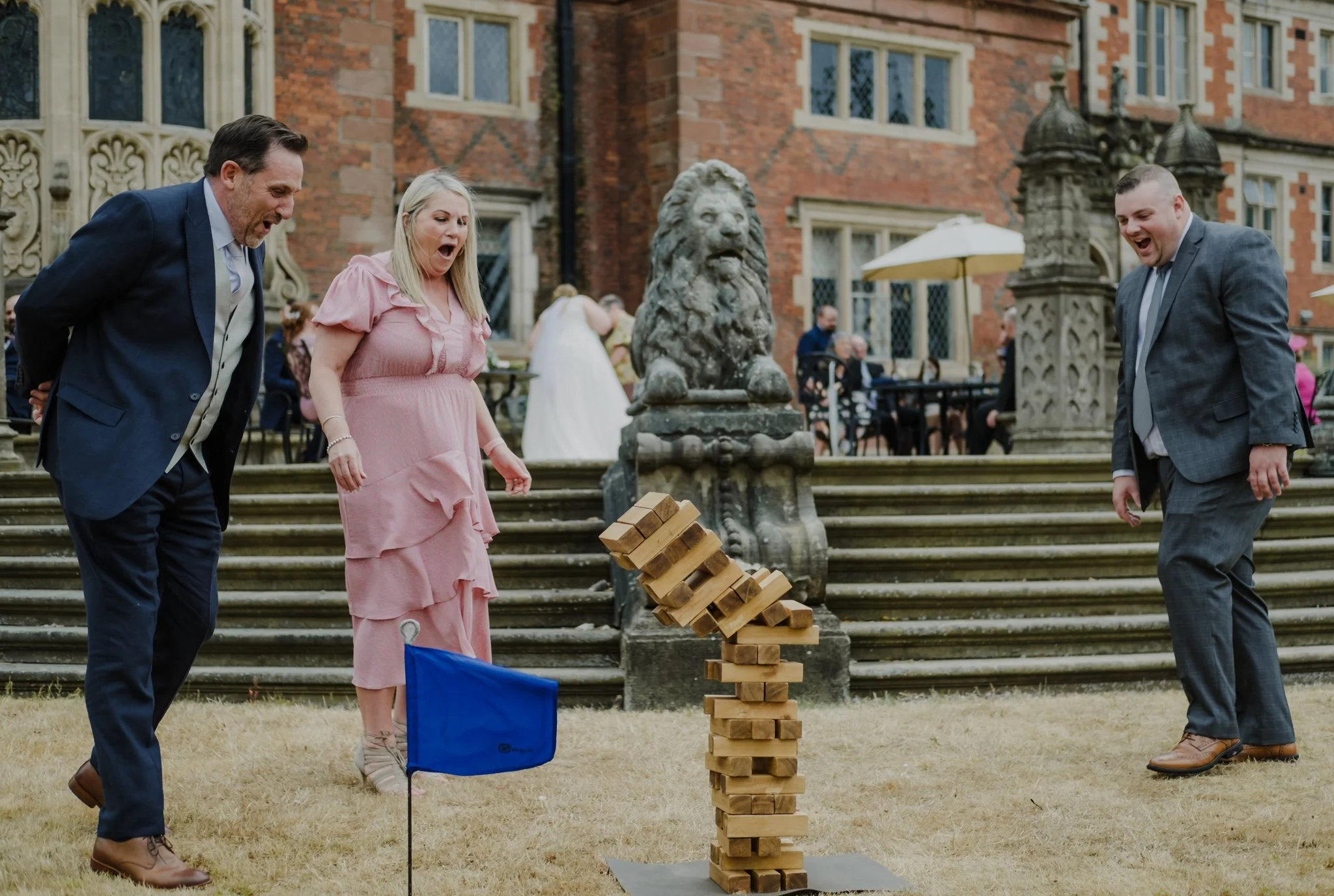 People playing giant outdoor Jenga game, with a woman in pink dress and two men in suits looking surprised, on the grass in front of a historic brick building with lion sculpture and stairs.