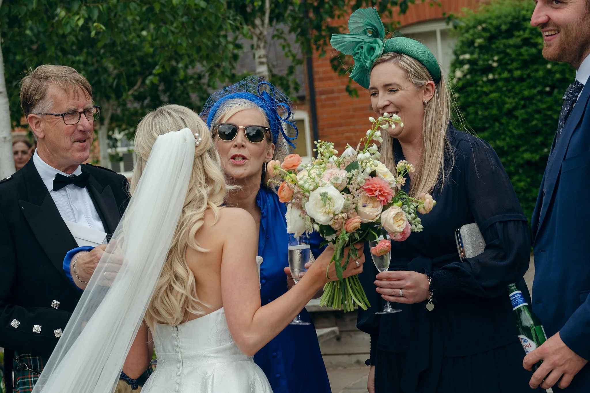 A bride in a white wedding dress with a veil and blonde hair is holding a bouquet of flowers and sharing a moment with guests at her wedding reception. Four adults, including a woman with sunglasses and a blue fascinator, are gathered around her, smi