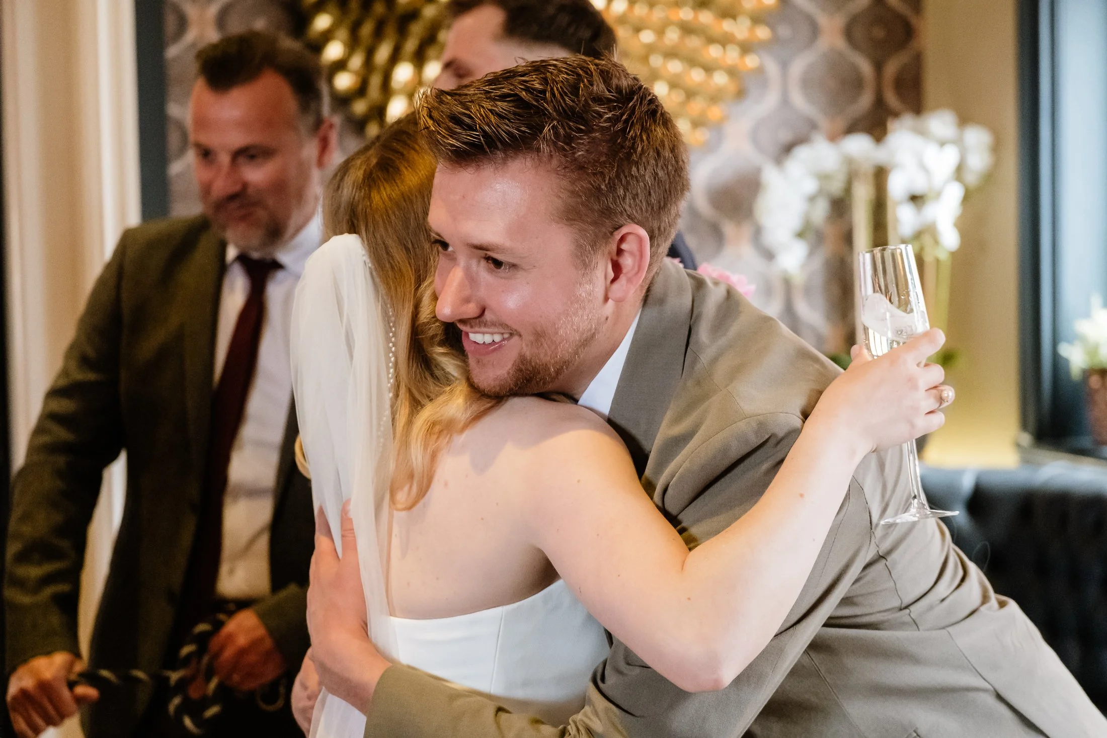A joyful man in a tan suit hugging a woman in a white dress at a celebration, holding a glass of champagne, with other people in the background.