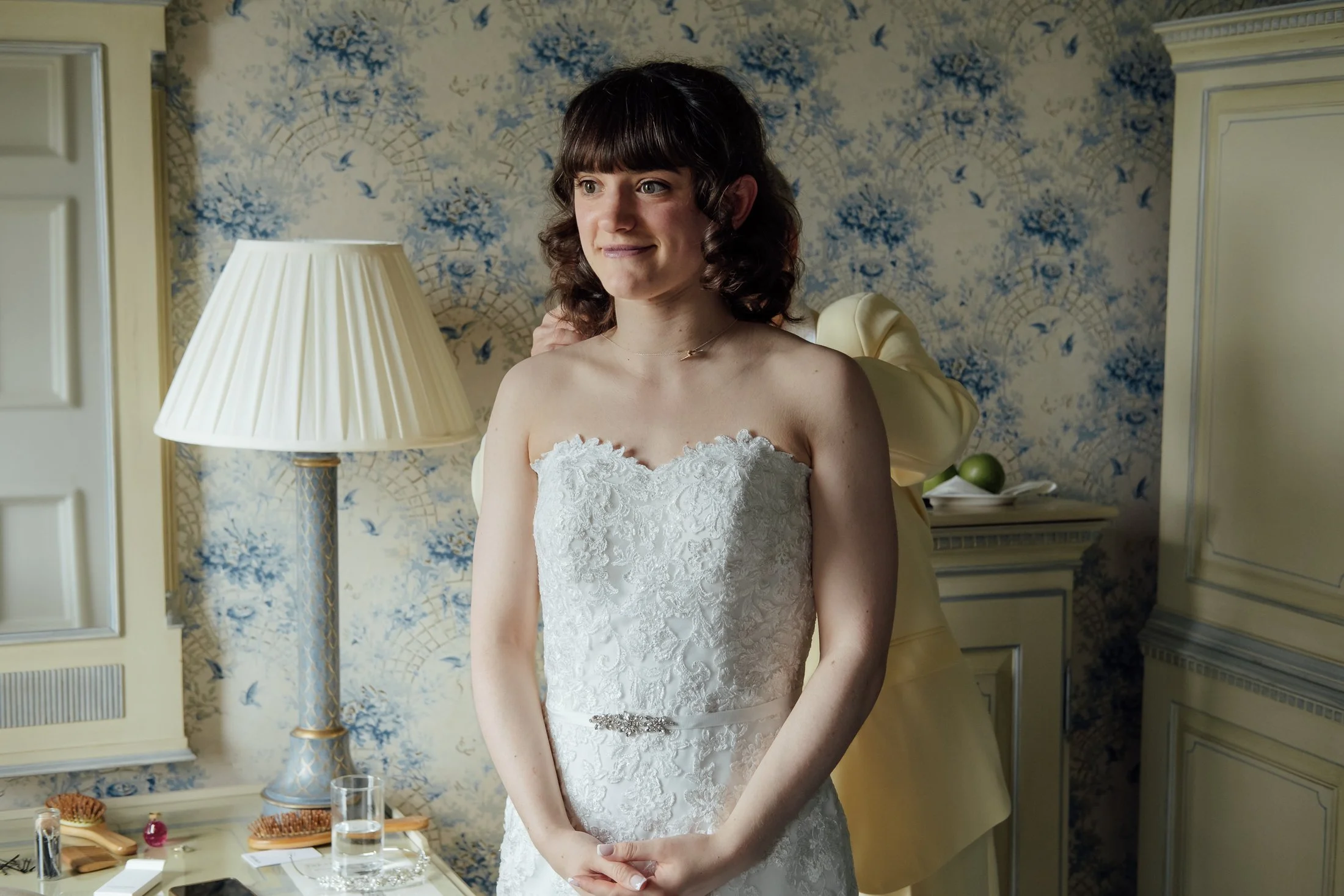 A woman in a wedding dress standing in a room with floral patterned wallpaper and a table with toiletries. She is smiling and looking to the side, with a person helping her in the background.