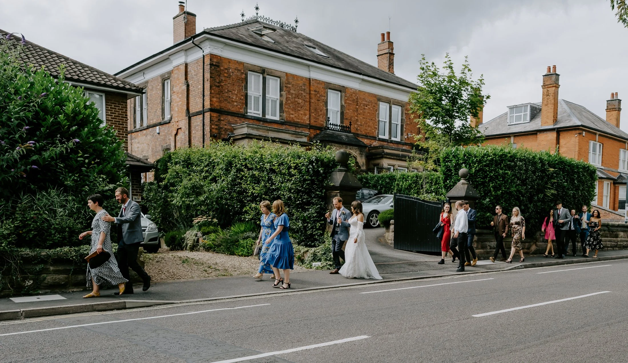 A group of people in formal attire walking on the sidewalk in front of a large brick house with greenery and parked cars.